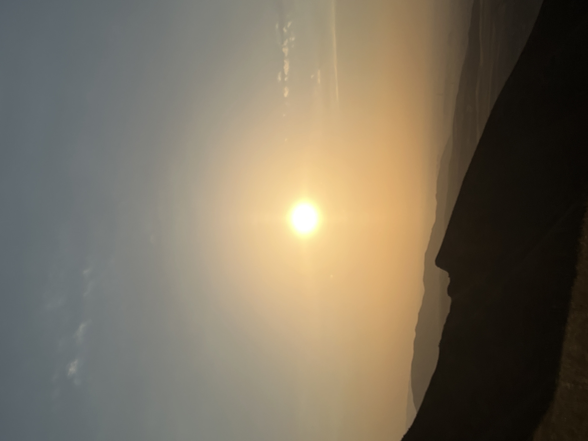 The image captures a sunset viewed from a mountain overlooking the city of Bishkek, Kyrgyzstan. The sun is a bright, intense orb positioned slightly left of the center of the frame, casting a warm, golden-orange glow across the sky. The sky transitions from a pale blue at the top to a hazy, yellowish-orange near the horizon. Faint wisps of clouds are visible, catching the light.
In the foreground, the dark silhouette of a mountain slope dominates the bottom left portion of the image, with dry, yellowish grass visible at the very bottom. The middle ground is filled with layers of silhouetted mountain ranges receding into the hazy distance, creating a sense of depth. These mountains are dark and indistinct, softened by the atmospheric conditions and the fading light. The overall impression is one of vastness and natural beauty, with the distant city of Bishkek likely obscured by the haze and the mountain ranges.