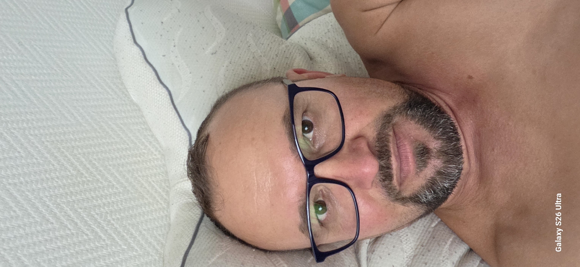 A man with short, receding brown hair and a goatee is lying in bed, looking at the camera. He is wearing blue-rimmed glasses. The bed appears to be in a bedroom in a home. The background is a white textured surface, possibly a headboard or wall. A portion of a pillow with a blue and white checkered pattern is visible on the right side of the image. The lighting suggests a natural light source, possibly from a window. The text "Galaxy S26 Ultra" is visible in the bottom left corner of the image. No specific location in the city of Evergem, Belgium is depicted.