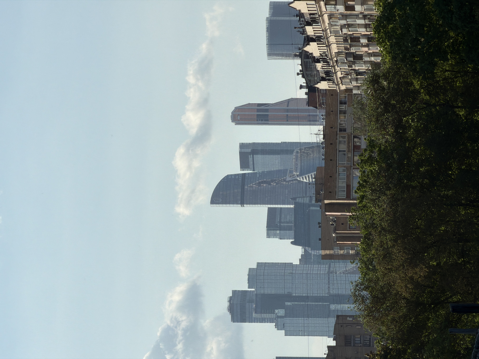 The image shows a view of the Moscow International Business Center (Moscow City) skyline, with modern skyscrapers rising above older residential buildings and trees. The sky is light blue with some clouds. The image appears to be taken from a lower vantage point, looking up at the skyscrapers, with foreground trees partially obscuring the view.