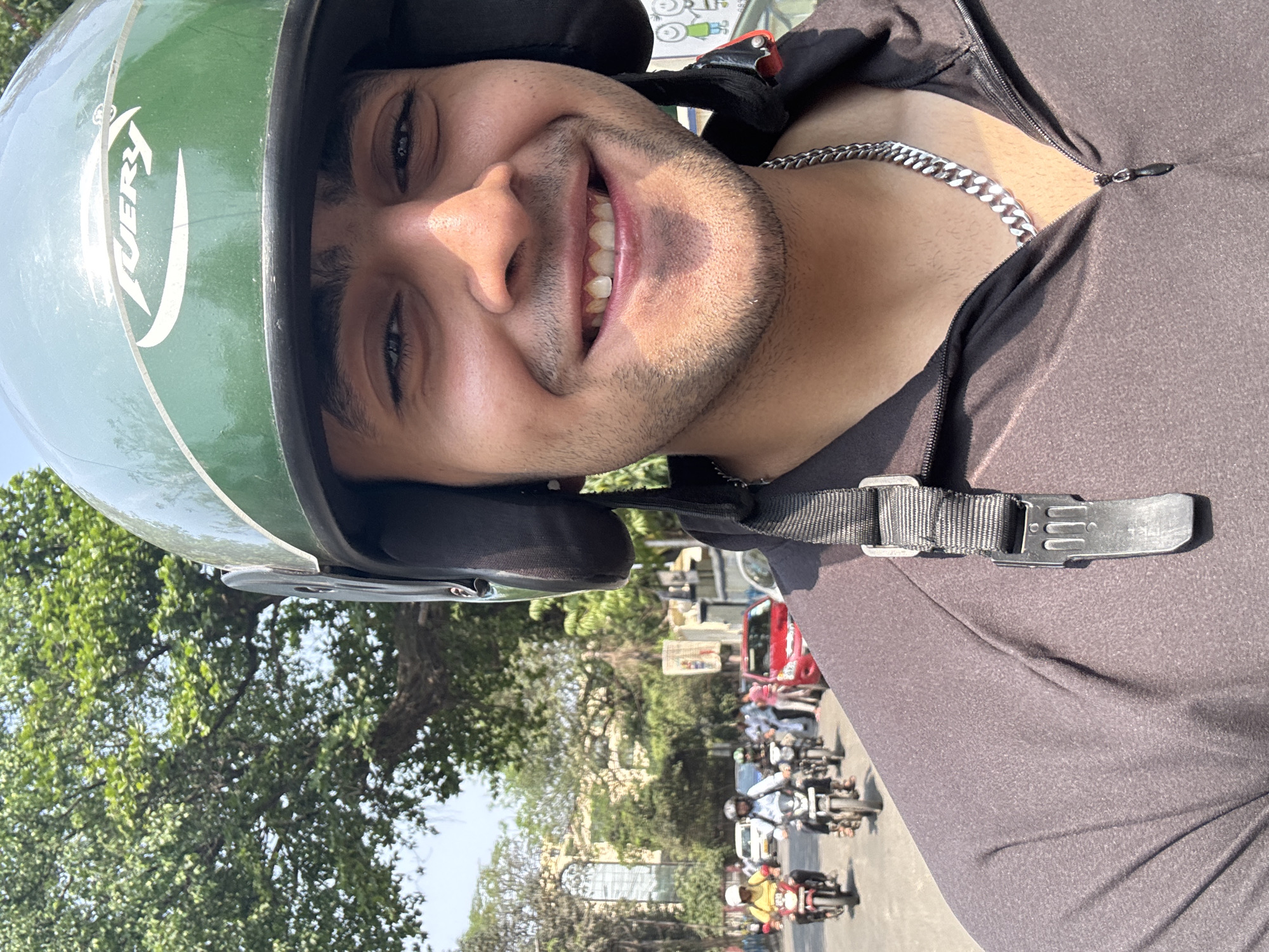 A young man wearing a green "VERY" branded motorcycle helmet is taking a selfie while riding a motorcycle on a busy street in Kolkata, India. He is wearing a dark grey zip-up jacket and a silver chain. The street is crowded with other motorcycles, pedestrians, and vehicles. The background features trees, buildings, and a clear sky.
