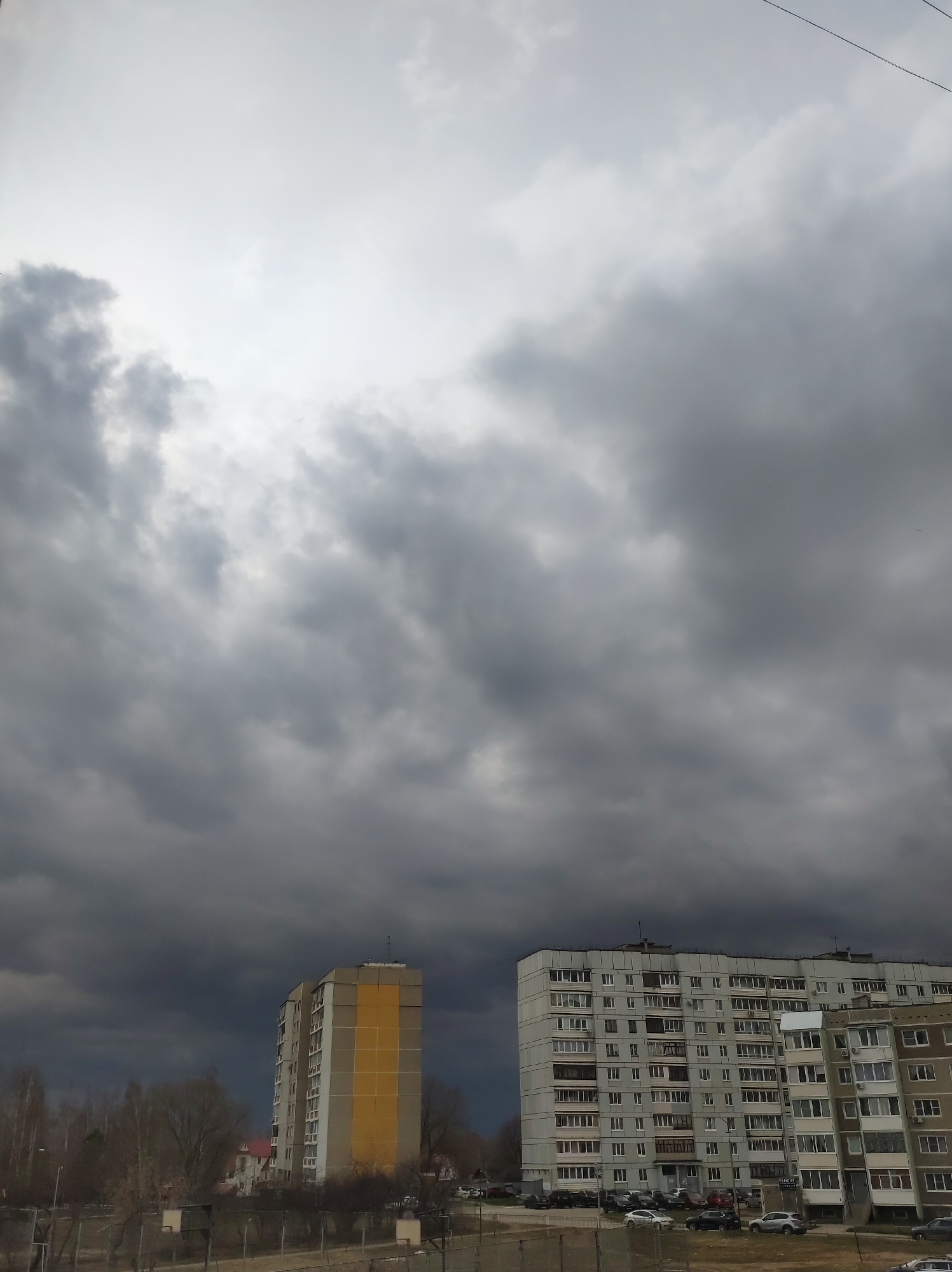 The image shows a cloudy sky over a residential area in Chaikovsky, Russia. Two multi-story apartment buildings are visible, one with a section painted yellow. Several cars are parked near the buildings. Trees and a fence are also visible in the foreground. The sky is filled with dark, ominous clouds, suggesting an approaching storm.
