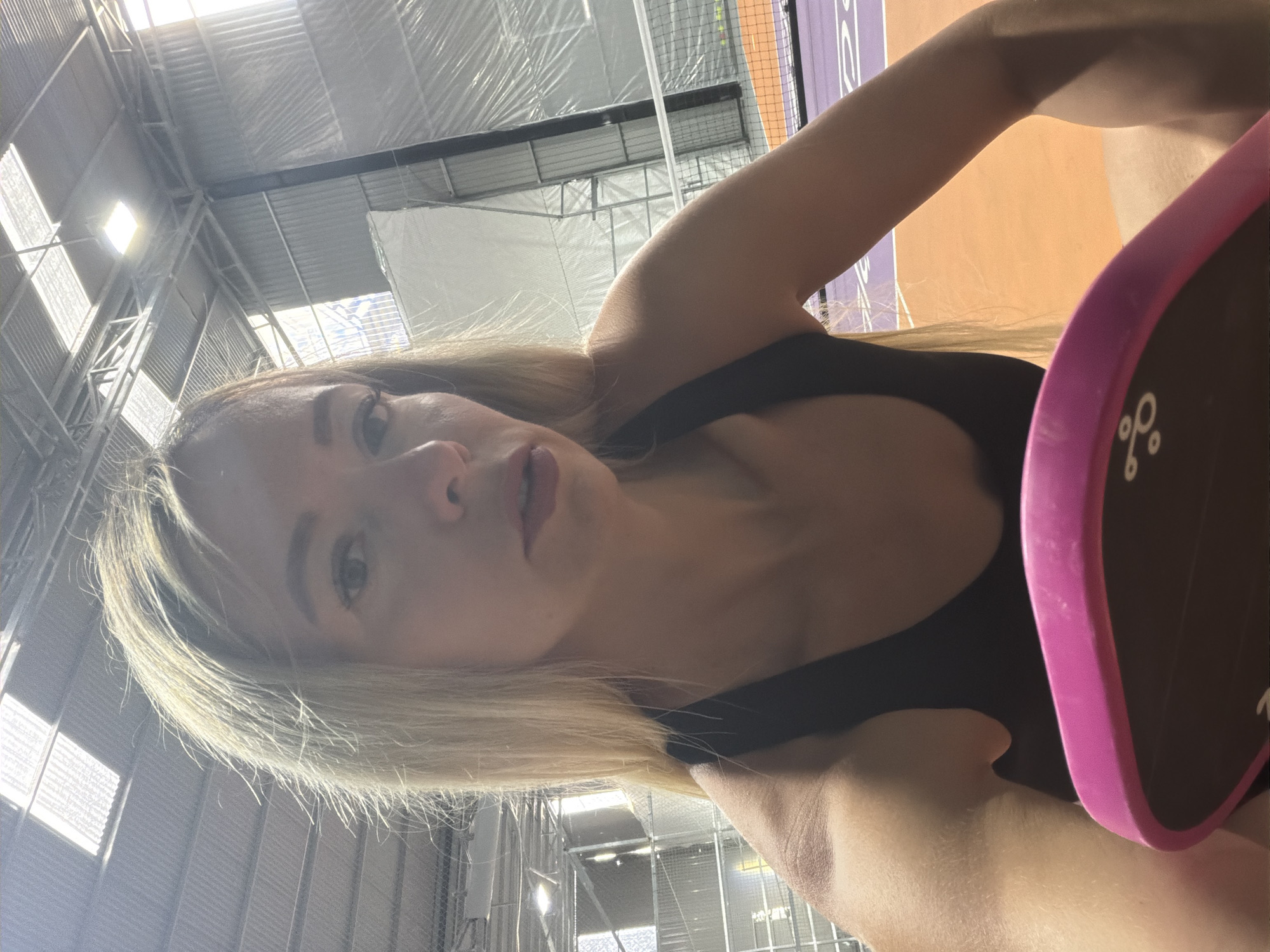 In a bright indoor sports facility, likely a pickleball or tennis court due to the net and court markings visible in the background, a blonde woman is taking a selfie. She is wearing a black athletic top and her arm is visible in the foreground, holding a pink pickleball paddle. The environment appears to be a covered court with a metal roof structure and large windows allowing natural light to filter in, creating bright highlights. The court surface is orange and purple.