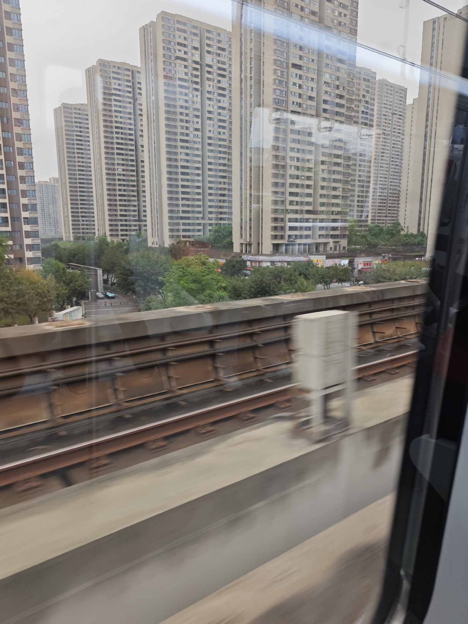 A view from a moving train in Chongqing High-tech Zone, China. Outside, rows of tall residential buildings dominate the skyline. A lower concrete barrier runs parallel to the train tracks. Between the barrier and the buildings are trees, roads, and cars. The photograph is taken through the train window.