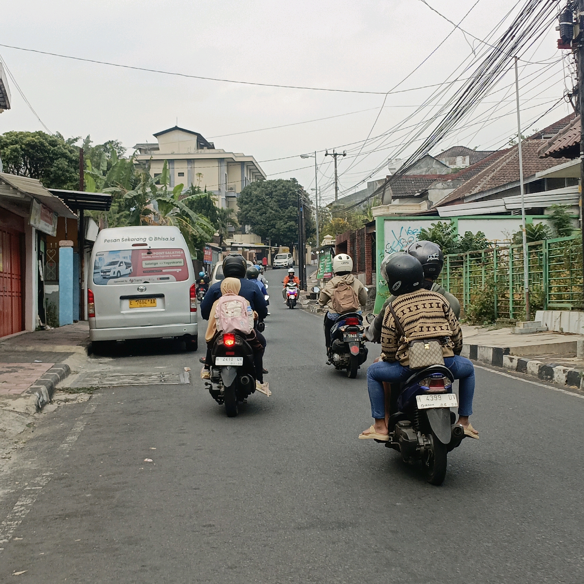 The image captures a street scene in Salatiga, Indonesia, where several motorcycles are navigating the road. In the foreground, a motorcycle carries two individuals; the rear passenger, a woman wearing a brown and black patterned long-sleeved shirt and blue jeans, has a small beige handbag slung across her body. The driver of this motorcycle is partially obscured. To the left, another motorcycle is visible with two passengers, one of whom is a child wearing a pink backpack with cartoon images. Further down the road, more motorcycles, including one carrying a rider in an orange jacket, are moving in the same direction. A silver van with advertisements on its rear is parked or moving on the left side of the street. The environment appears to be a mixed urban area with buildings of varying sizes, including what seems to be an apartment complex or commercial building in the background. Utility poles with a dense network of electrical wires are prominent overhead, suggesting a developed urban setting. The road surface is asphalt, and there are sidewalks with curbs on the left. The sky is overcast.