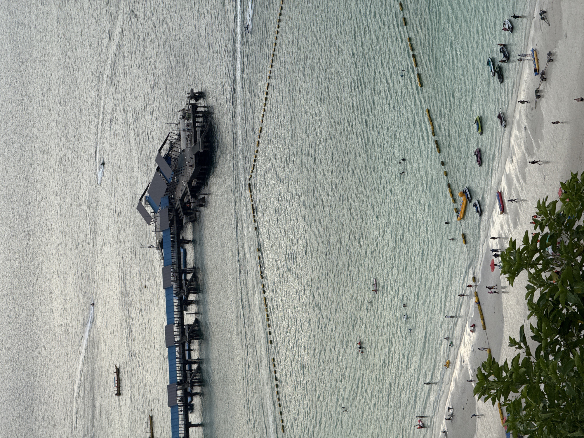 The image shows an aerial view of a beach in Pattaya City, Thailand. A pier extends into the water from the left side of the image. A line of buoys runs parallel to the beach. The beach is sandy and populated with people, including swimmers, sunbathers, and pedestrians. Several watercraft are visible on the beach, including boats and jet skis. Some green foliage is visible in the lower right corner. The water is light blue, and the sky is overcast.