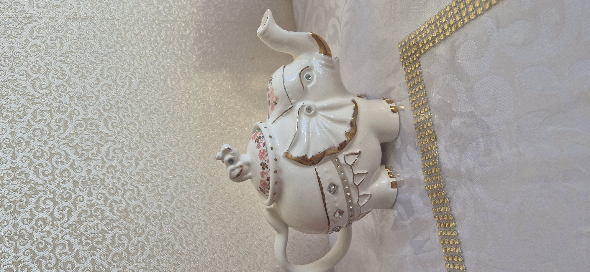 The image shows a white, ceramic elephant figurine, adorned with gold trim and floral patterns. It features bejeweled eyes and embellishments. The elephant is positioned against a textured, patterned wall or fabric, with a gold-colored, rhinestone-like border visible on the right side. The backdrop is primarily light-colored, possibly a wall or tablecloth, with an intricate design. The location and city are unknown.