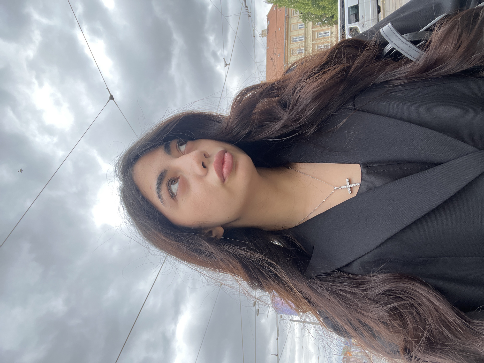 The image shows a young woman from the chest up, looking to her right. She has long, dark, wavy hair and is wearing a black blazer over a dark top and a silver cross necklace. The background appears to be an urban environment with a cloudy sky, overhead wires, and buildings in the distance. The overall impression is of a candid outdoor shot. The location of the image has been identified as Pilsen, Czechia.
