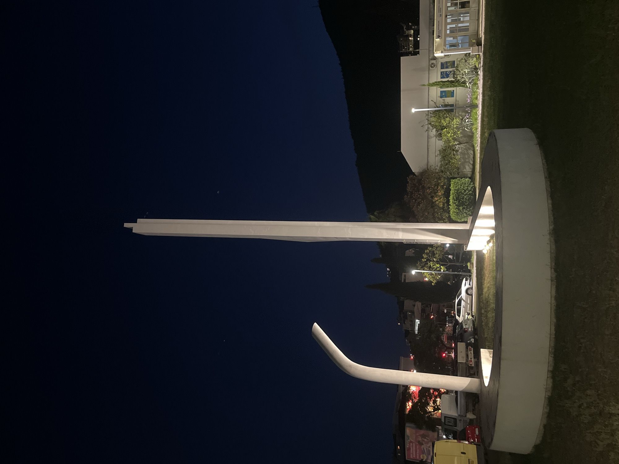 An eye-level, medium outdoor shot captures a modern monument at night in an unknown city. The monument is a white, abstract sculpture featuring a tall, slender vertical element with a slight curve at the top, and a large, curved base that creates a circular enclosure. The sculpture is illuminated from within the base, casting a warm glow.
In the background, a dark, tree-covered hillside rises against a deep navy blue night sky. A low-rise building with a white facade is visible to the right of the monument, featuring windows and an entranceway. The building appears to be an educational or public institution, as indicated by text on the facade and colorful artwork visible through some windows.
Several cars are parked in front of the building and on a street to the left of the monument. A yellow van is partially visible on the far left. Trees and landscaping surround the monument and the building, with some of the foliage illuminated by artificial lights. A streetlamp is visible near the building. The overall scene suggests a quiet, urban environment at night, with the monument serving as a central focal point.