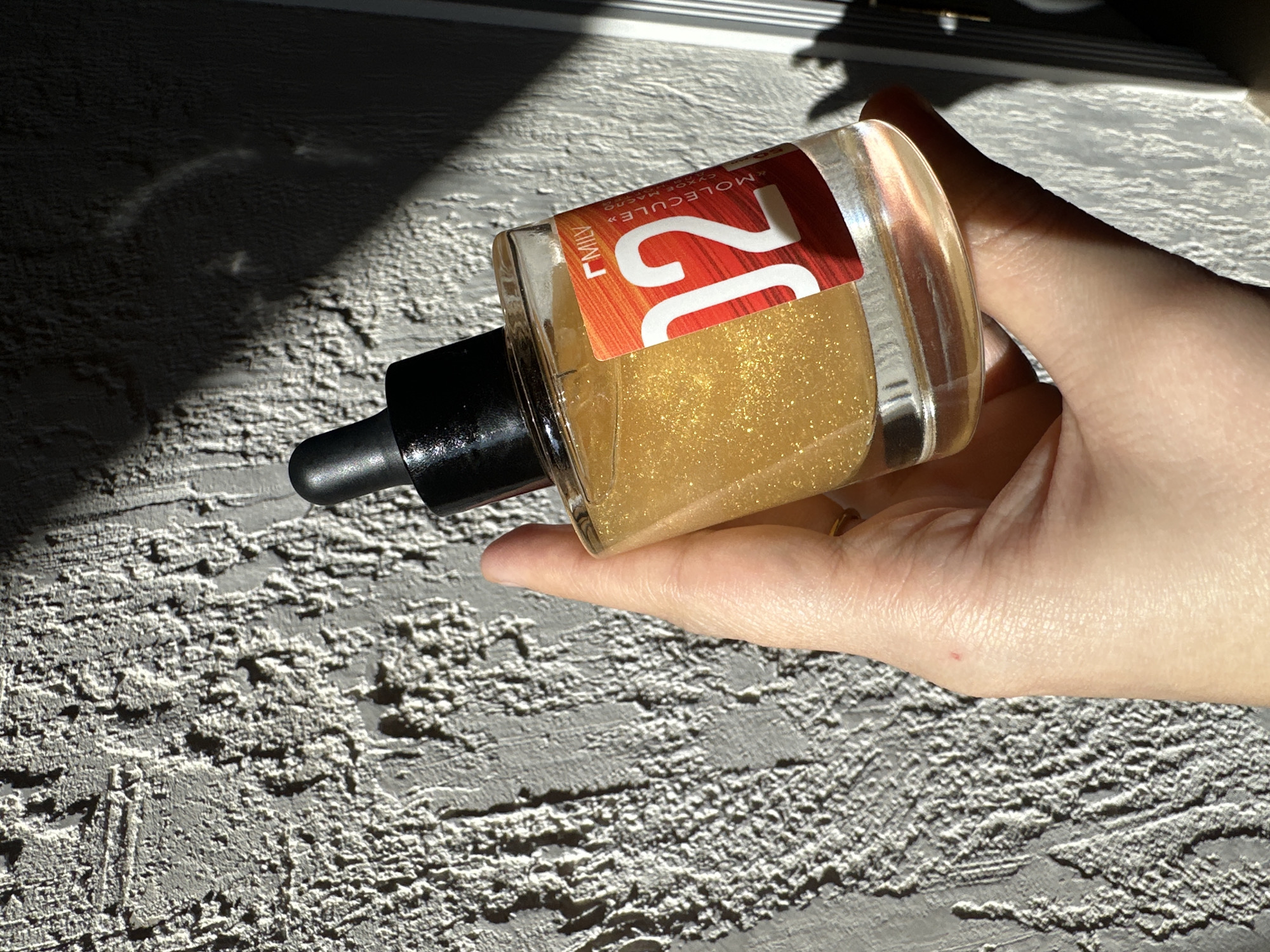 A hand holds a bottle of "FAMILY 12 MOLECULE+" liquid on a textured gray surface. The bottle is rectangular with a black dropper and a label with the number "12" in bold red and orange. The liquid inside appears golden and shimmering. The background is partially obscured by shadow and a metallic object. The location in Kharnavy, Russia is not determinable from the image.