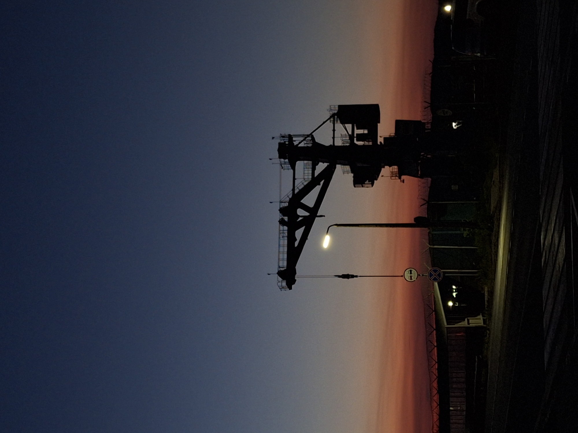 The image depicts a dark, silhouetted industrial crane against a twilight sky in the city of Chaikovsky, Russia. The sky transitions from a deep blue at the top to hues of orange and red near the horizon, indicating either sunrise or sunset. A single, illuminated street lamp is positioned to the right of the crane. Two circular traffic signs are visible on a pole near the street lamp, one with a red border and a white center, and another with a blue center and a red diagonal cross. In the background, a fence with barbed wire runs horizontally, and beyond that, there is a dark, indistinct landscape. A portion of a building or structure with lighting is visible behind the fence. The foreground shows a dark, paved area, possibly a road or pathway, with some textured lines that could indicate a pedestrian crossing. A vehicle is partially visible on the far right, also in silhouette. The overall scene suggests an industrial or port area in Chaikovsky during low light conditions.