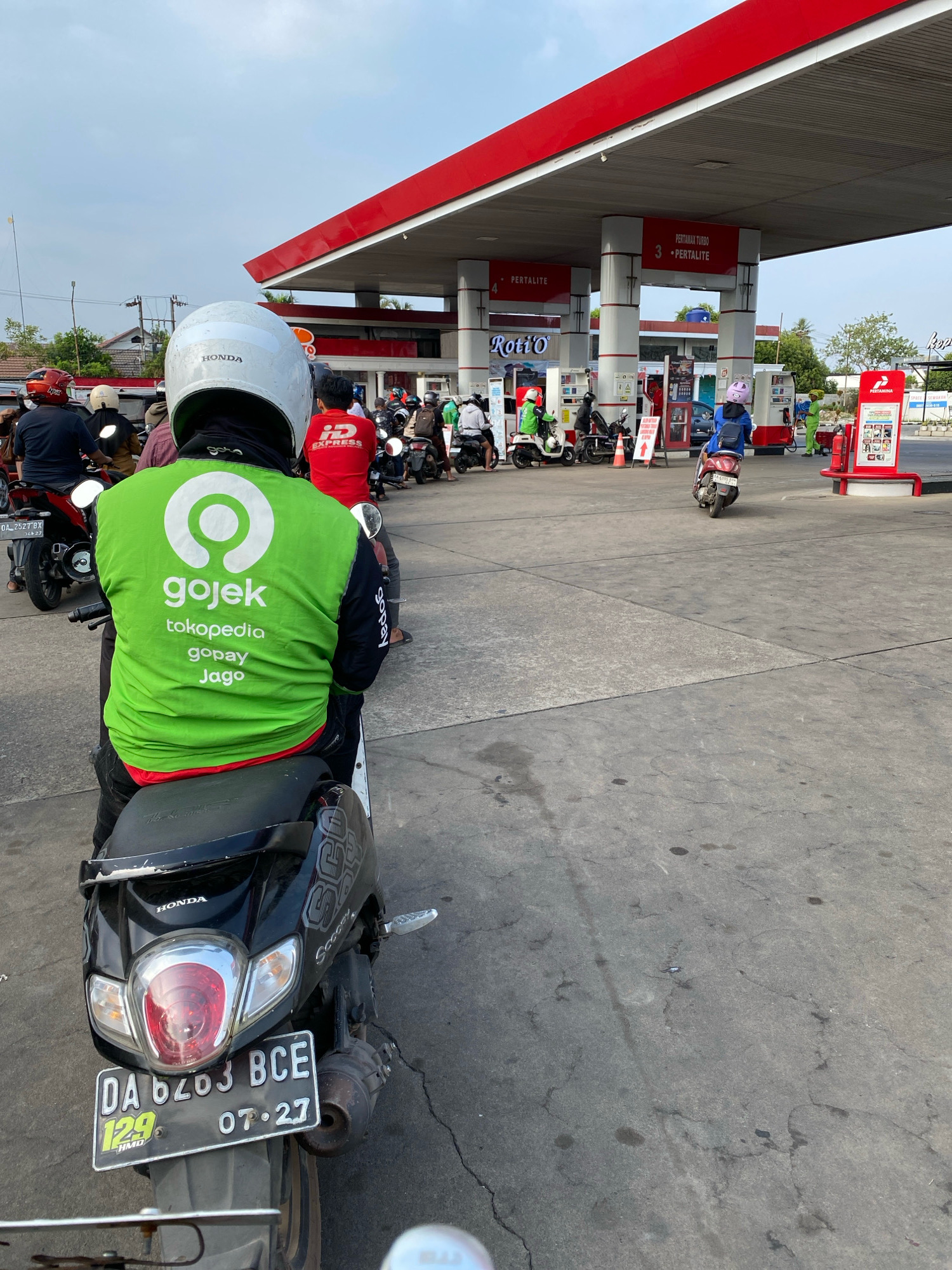 In Banjarbaru, Indonesia, a group of motorcyclists are lined up at a Pertamina gas station. The foreground features a person wearing a green Gojek jacket and a white helmet, seated on a black Honda scooter with license plate DA 6703 BCE. The scooter is positioned on a cracked concrete surface. Behind this individual, other people on motorcycles are waiting in line, some wearing helmets and jackets.
The gas station has a red and white canopy supported by pillars. Signs for "Pertamax Turbo" and "Pertaminte" are visible. A "Roti'O" sign is also present. In the background, more vehicles and individuals are present, with buildings and trees further in the distance. The sky is overcast. The scene depicts a common occurrence of fuel queues at a gas station.