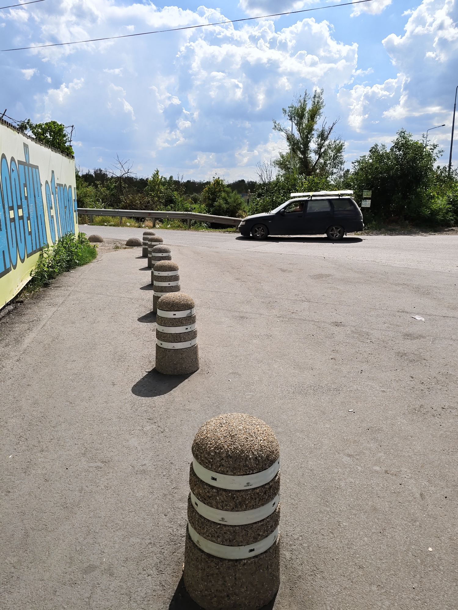 A street scene in Rostov-on-Don, Russia, features a row of concrete bollards with reflective white bands lining the edge of an asphalt road. The bollards are positioned on the right side of the foreground, leading the eye towards the background where a dark-colored station wagon is driving away. To the left, a graffiti-covered wall with blue lettering partially obscures a metal guardrail. Lush green vegetation and trees are visible beyond the guardrail, and the sky is filled with scattered white clouds against a blue backdrop. The overall environment suggests an outdoor setting, possibly a roadside or an industrial area.