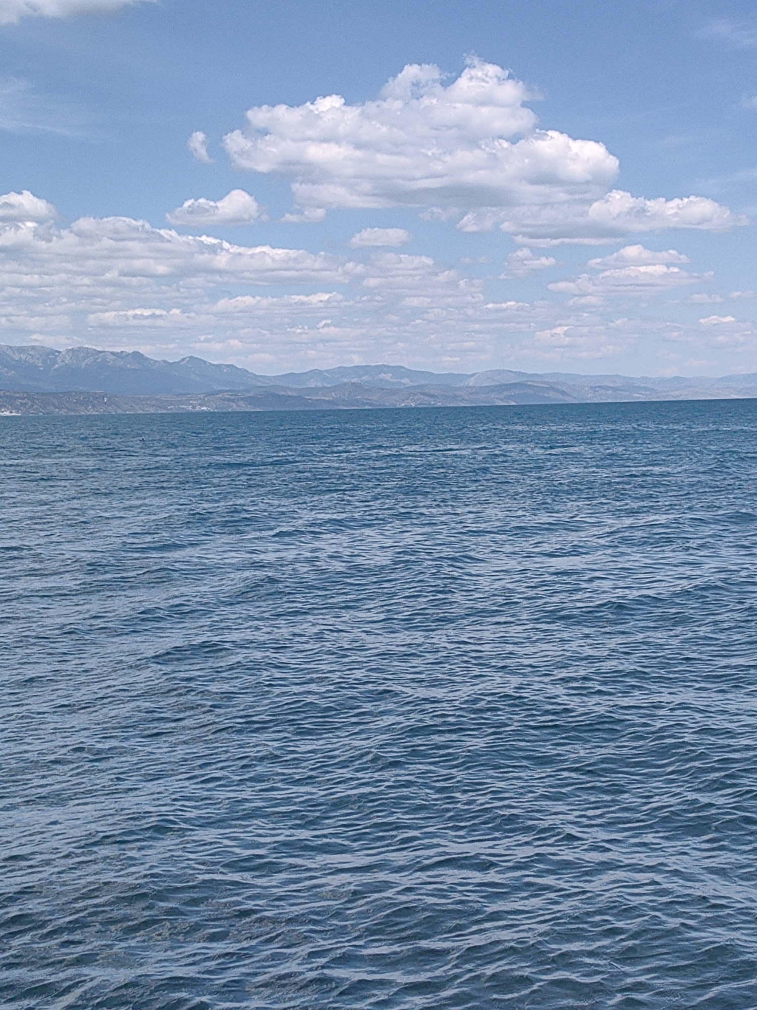 The image shows a wide expanse of blue water with small waves, stretching towards a distant mountain range under a partly cloudy sky. The mountains are hazy and appear to be a light blue or gray color. The sky is a bright blue with scattered white cumulus clouds. There are no visible people, structures, or immediate signs of human activity in the scene. The overall impression is a natural landscape, likely a large body of water such as a lake or sea, with mountains in the background. The location in Utios, Ukraine, suggests this could be a view of the Black Sea coast or a large inland body of water in that region.