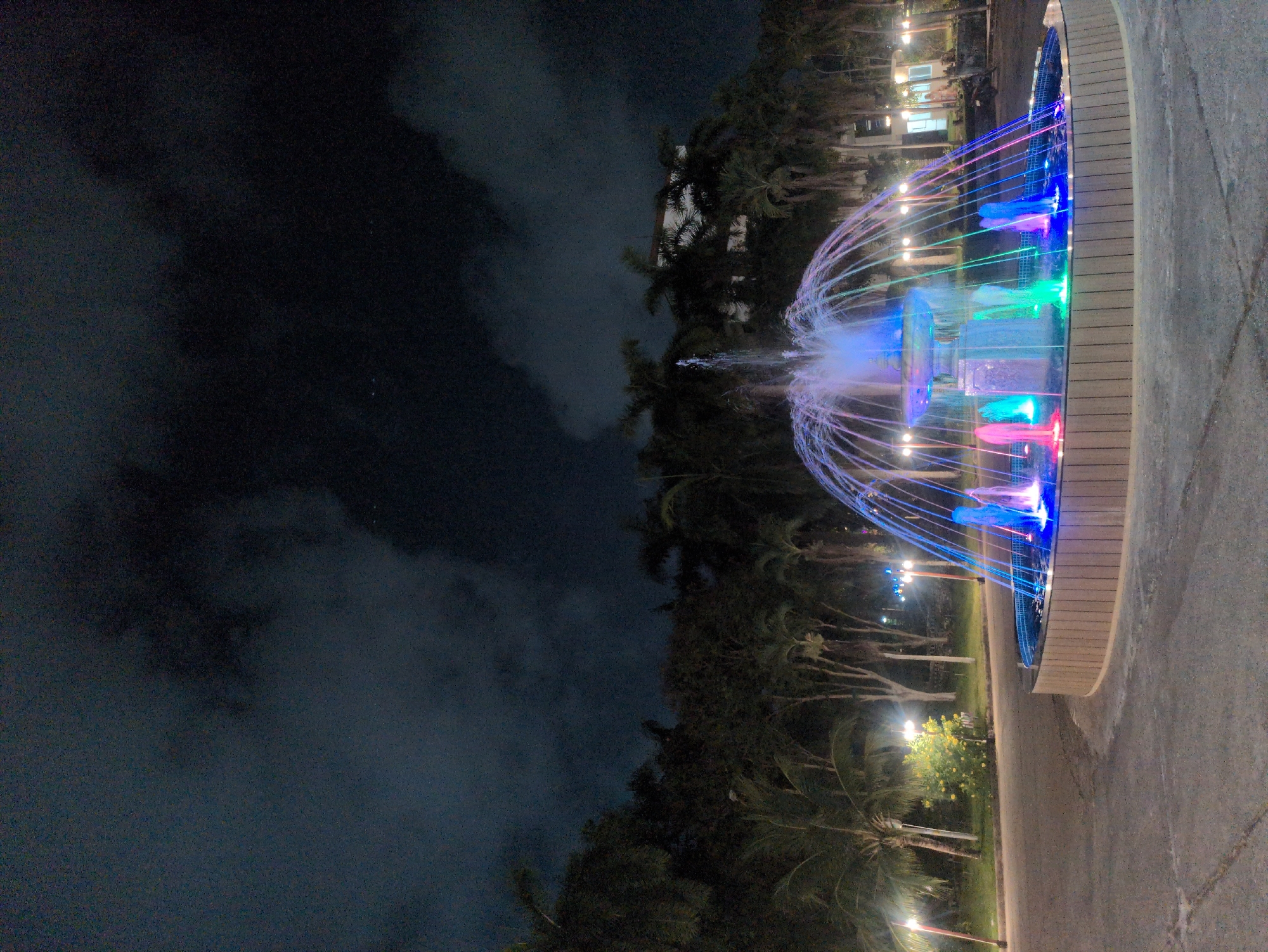 A nighttime scene shows a brightly lit, tiered fountain in a park in Phú Quốc, Vietnam. The fountain is circular, lined with a light-colored, vertically ridged border. Water streams arc upward from the fountain, illuminated with vibrant blue, green, and pink lights. A paved walkway extends from the fountain. In the background, palm trees and other vegetation are visible, some illuminated by streetlights. Beyond the trees, the night sky is dark, with clouds obscuring any stars.