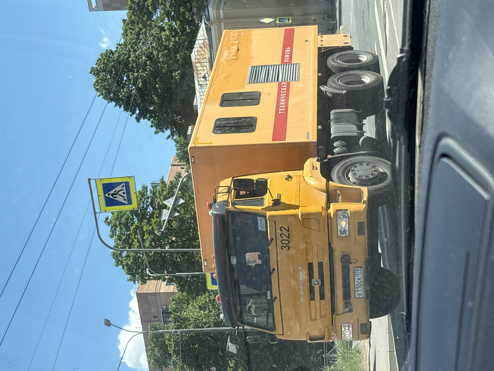 This is a close-up, low-angle shot of a yellow Kamaz truck driving down a street in Saint Petersburg, Russia, on a sunny day. The truck has a large cargo box on its back, which is also yellow and has red stripes. Text on the side of the cargo box reads "ТЕХНИЧЕСКАЯ ПОМОЩЬ" (Technical Assistance). The truck's license plate reads "X110CK 178".
In the background, there are trees, buildings, and a pedestrian crossing sign. The sky is blue with a few clouds. The overall impression is a typical urban scene in Russia. The time of day appears to be midday, given the bright sunlight and clear sky. There are no people clearly visible in the image, but the presence of the truck suggests it is either operating or parked. The setting appears to be a commercial or industrial area, as indicated by the type of truck and the surrounding infrastructure. The camera is positioned inside another vehicle, looking out at the truck.