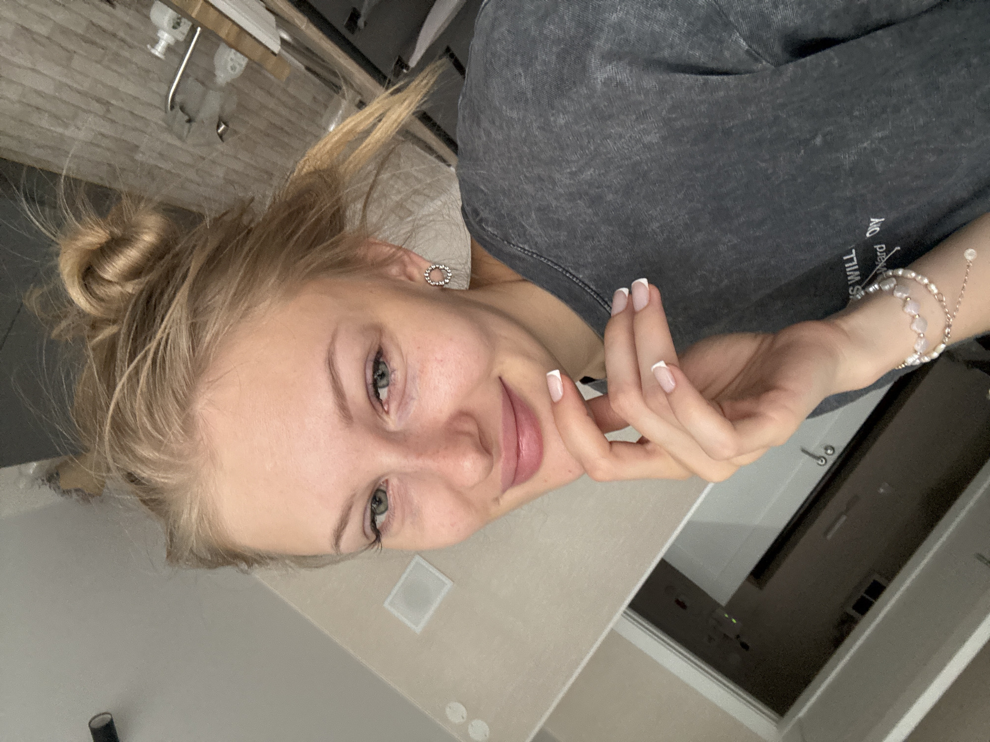A young woman with blonde hair styled in a bun and green eyes is captured in a close-up, head-and-shoulders selfie. She is wearing a dark gray t-shirt and a bracelet on her left wrist, which consists of pearls and pink heart-shaped beads. Her nails are manicured with a French tip. The woman is resting her chin on her right hand, and a silver circular earring is visible on her right ear. She is indoors, with a wall in the background that appears to be painted a beige color. A doorway is visible to the left of the frame, leading to a darker room. The image appears to be taken in a residential setting. There is no indication in the image that this location is the city of Perm, Russia.
