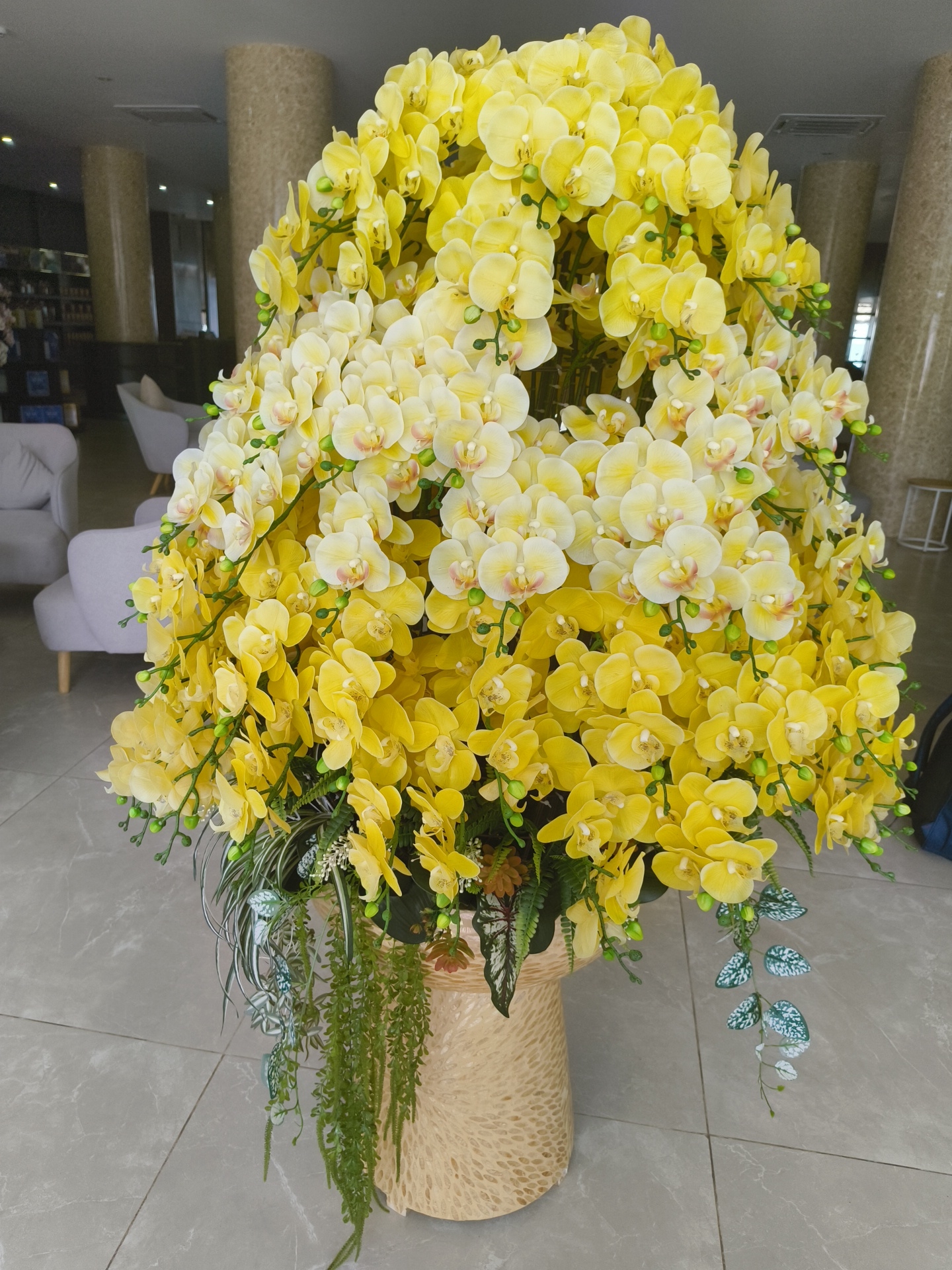 A large, artificial bouquet of yellow and cream orchids is displayed in a woven pot. The bouquet is dense and voluminous, with multiple stems of flowers and buds cascading downwards. Some green foliage and other artificial plants, including trailing vines with speckled leaves, are interspersed within the arrangement. The bouquet is placed on a grey tiled floor, likely in an indoor setting. In the blurred background, there are hints of an interior space with beige pillars, seating areas with light-colored chairs, and shelves that might contain books or decorative items. The overall impression is one of a decorative centerpiece in a lobby or common area. The location is described as Phu Thuy Ward, Vietnam.