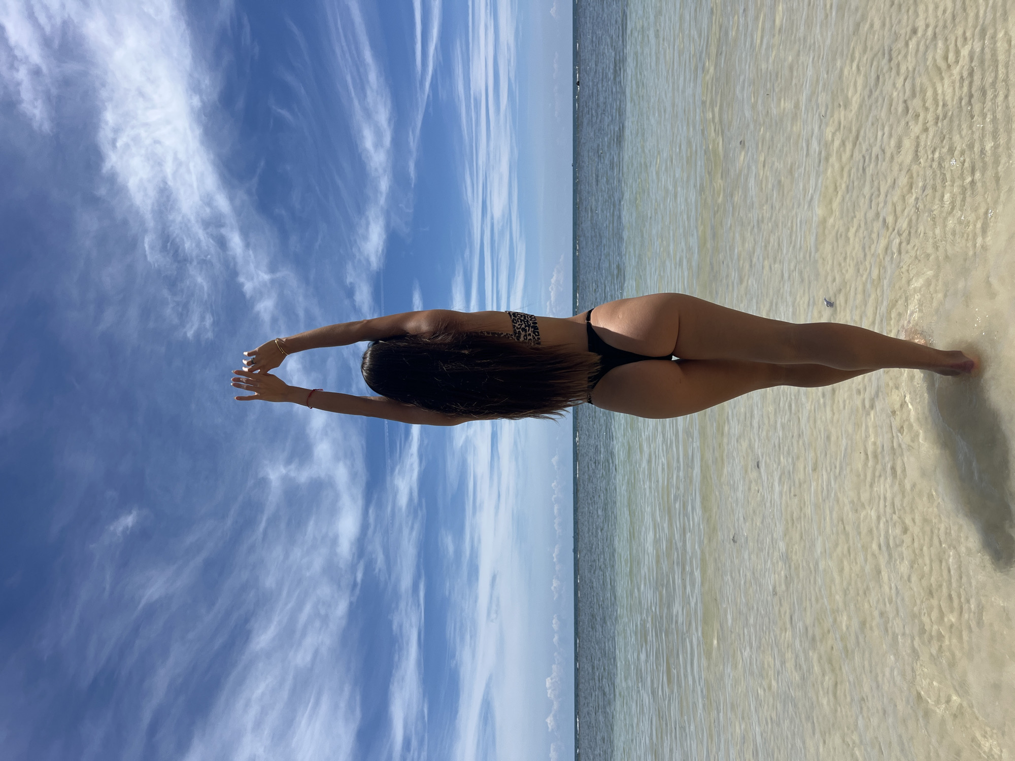 A rear view of a woman in a black bikini with a leopard print top stands in shallow, clear ocean water. Her arms are raised above her head, and her long, dark hair cascades down her back. The water is calm with a sandy bottom visible beneath the surface. In the background, the ocean meets the horizon under a bright blue sky filled with wispy white clouds. The location is identified as Koh Phangan, Thailand.