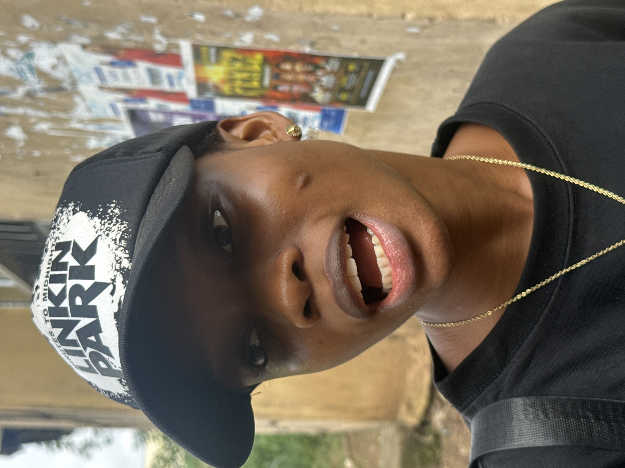The image shows a close-up portrait of a young Black person wearing a black baseball cap with "LINKIN PARK" printed on it and a black t-shirt. The person has a gold stud earring in their right ear and a gold chain necklace. Their mouth is open, revealing teeth and gums. The background consists of a rough, textured wall with posters and some greenery visible on the left. The setting appears to be outdoors in a part of Ihiagwa, Nigeria. The lighting suggests it is daytime.