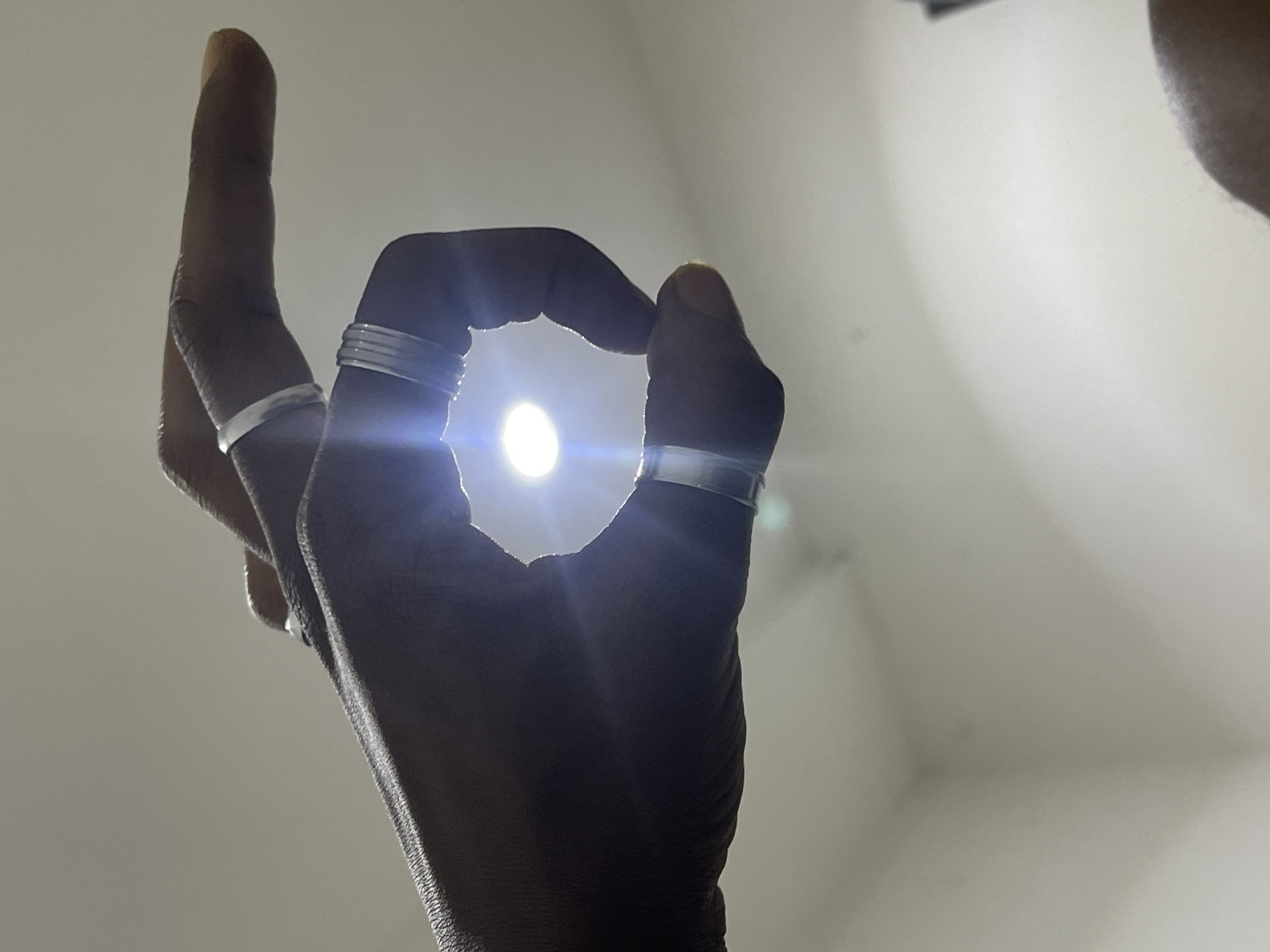 A low-angle, close-up shot shows a person's hands, which are adorned with several silver rings, held up against a brightly lit ceiling. The hand on the left is positioned to form a rough circle with its thumb and index finger. The hand on the right is extended, with its thumb and index finger also forming a circular shape. A bright light source, possibly a bulb or a flashlight, is visible through the gap between the fingers, creating a lens flare effect. The background consists of a plain, off-white ceiling with some subtle shadows, suggesting an indoor setting. The lighting is strong, casting shadows on the hands and highlighting the textures of the skin. No specific location in Abuja, Nigeria is identifiable from the image.