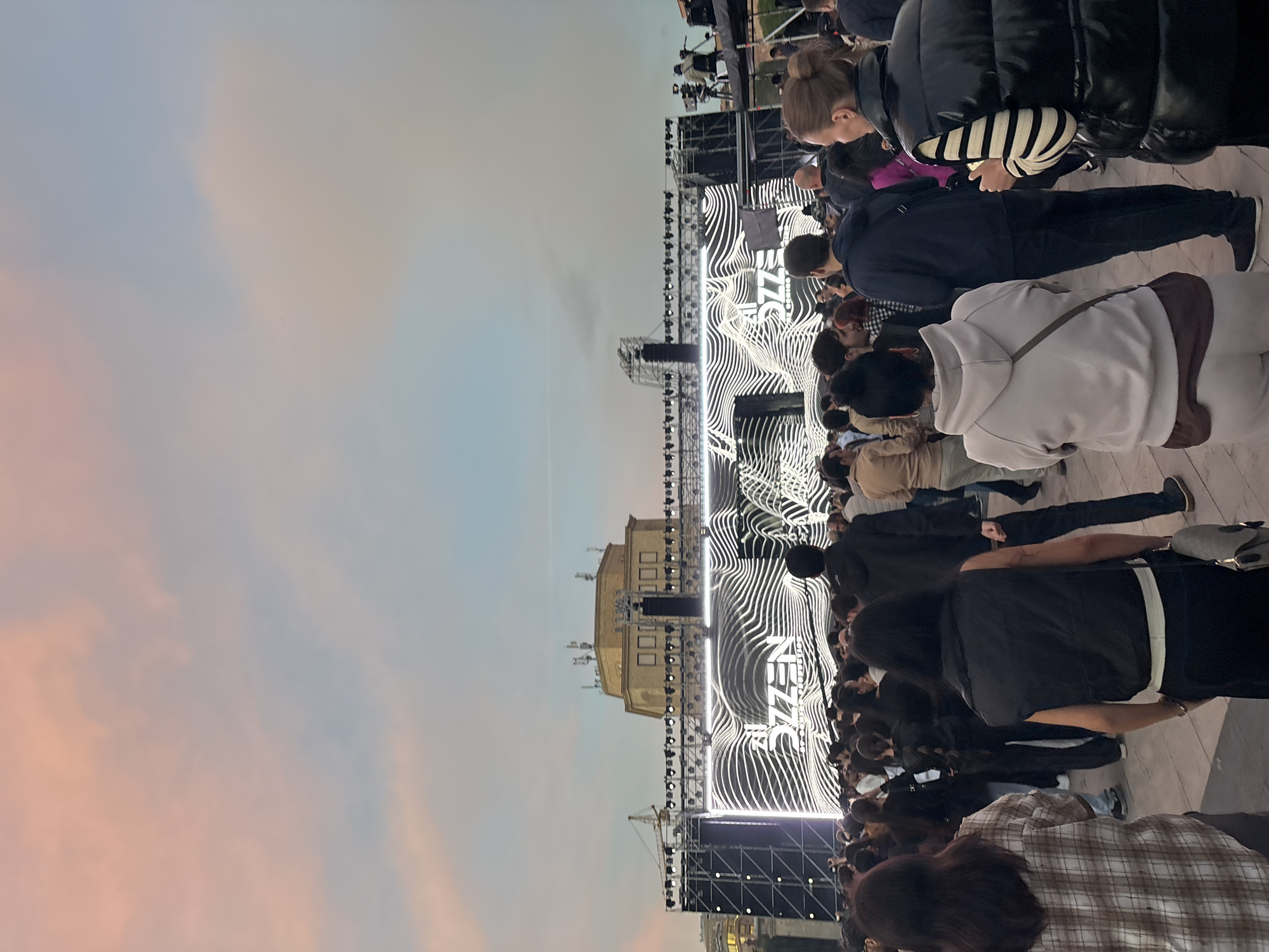 The image shows a crowd of people gathered at an outdoor event in front of a stage with a backdrop featuring a white, wavy design. The building in the background is likely in Italy. The sky is a mix of light blue, pink, and grey, indicating dawn or dusk. People are standing and facing the stage. Some are dressed in casual attire. There are lighting rigs and scaffolding surrounding the stage.