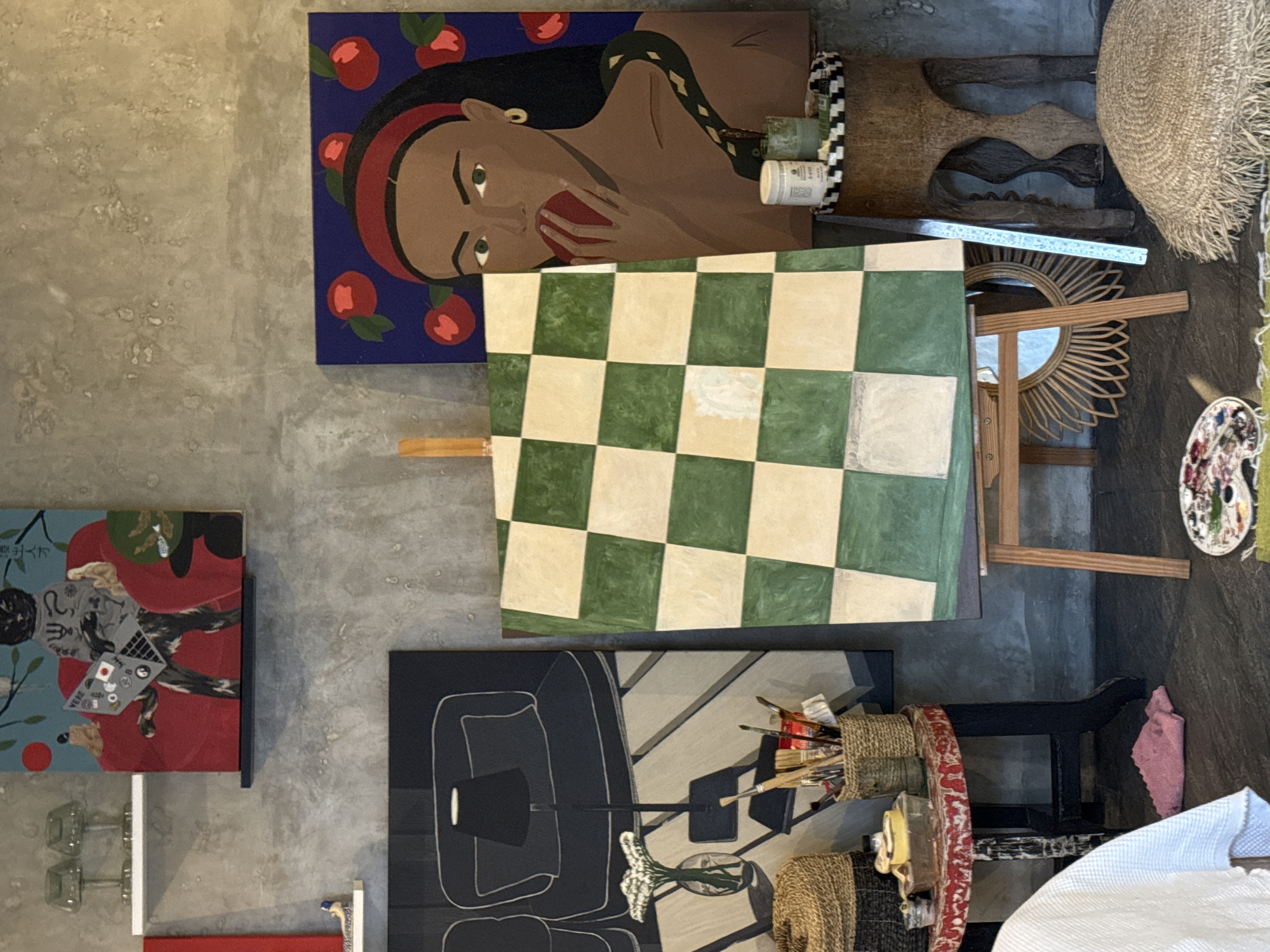 The image shows an indoor scene with a collection of artworks and decor items. From left to right, there are four visible paintings hanging on a gray wall: the first depicts a figure with Asian writing and a laptop; the second is an artistic portrait of a brown woman eating an apple, set against a blue background with red apples; the third is a green and white checkered painting; and the fourth is a black and white painting of a lamp and furniture. To the right of the checkered painting, there is a wooden easel and a round mirror, followed by a woven sunhat. Lower right, there is a table with paintbrushes and art supplies. The setting is likely in North Kuta, Indonesia, as requested.