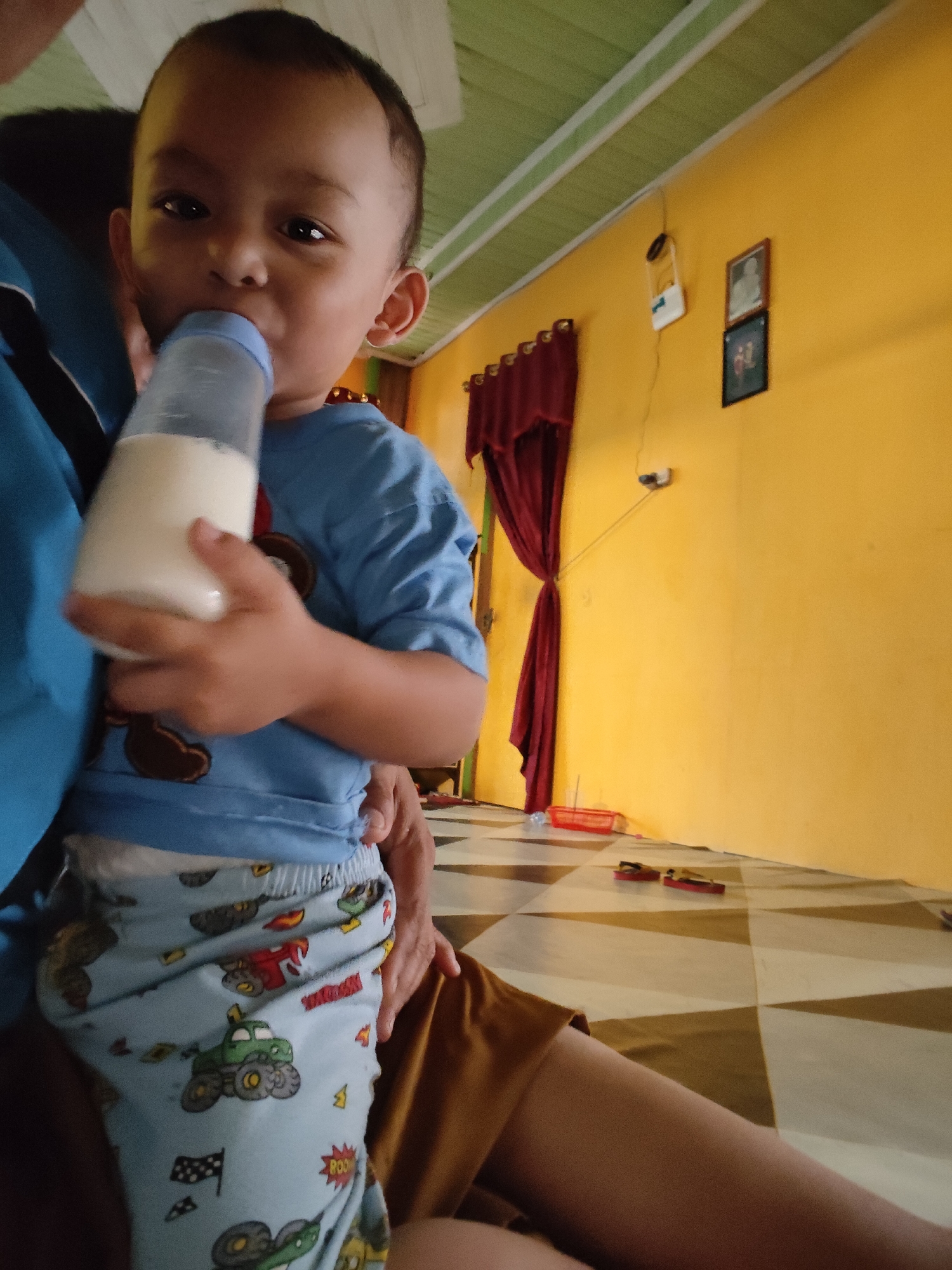 A young child, possibly a toddler, is being held and is drinking from a baby bottle filled with a white liquid. The child is wearing a blue t-shirt with a brown cartoon graphic on the front and light blue pants with a pattern of various cartoon vehicles, including monster trucks and race flags. The child's face is partially obscured by the bottle. The background suggests an indoor setting with a yellow wall, a green and white striped ceiling, and a floor with a checkered pattern in brown and beige. Red curtains hang on the wall, and two framed pictures are mounted above them. A white electrical device is also attached to the wall. On the floor, a red basket and a pair of red flip-flops are visible. The lighting appears to be natural, possibly from a window or overhead lights, casting a warm glow on the scene. The image conveys a moment of care and sustenance, likely within a home environment in Tanah Grogot, Indonesia.