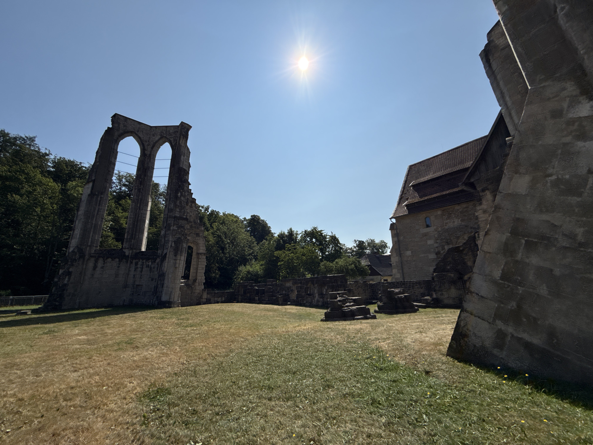 This is a wide-angle, exterior, eye-level shot of the Walkenried Abbey ruins in Walkenried, Germany, on a clear, sunny day. The sun is prominently featured in the upper center of the frame, casting a bright glare.
In the foreground, a grassy field dominates the lower half of the image, with patches of dry, golden grass interspersed with greener areas. Small yellow wildflowers are visible in the grass.
To the left, a large, crumbling stone structure with two prominent arched openings stands against a backdrop of lush green trees. This structure appears to be the remnants of a significant part of the abbey, possibly a nave or a tower.
To the right and center, another stone building, partially intact and appearing to be a more preserved section of the abbey, is visible. This building has a pitched, tiled roof. In front of this building and further into the background, there are various low-lying stone ruins and foundations scattered across the field.
The overall scene depicts the historical remnants of the Walkenried Abbey, showcasing its architectural past against a bright, natural outdoor environment. There are no people or animals present in the image.