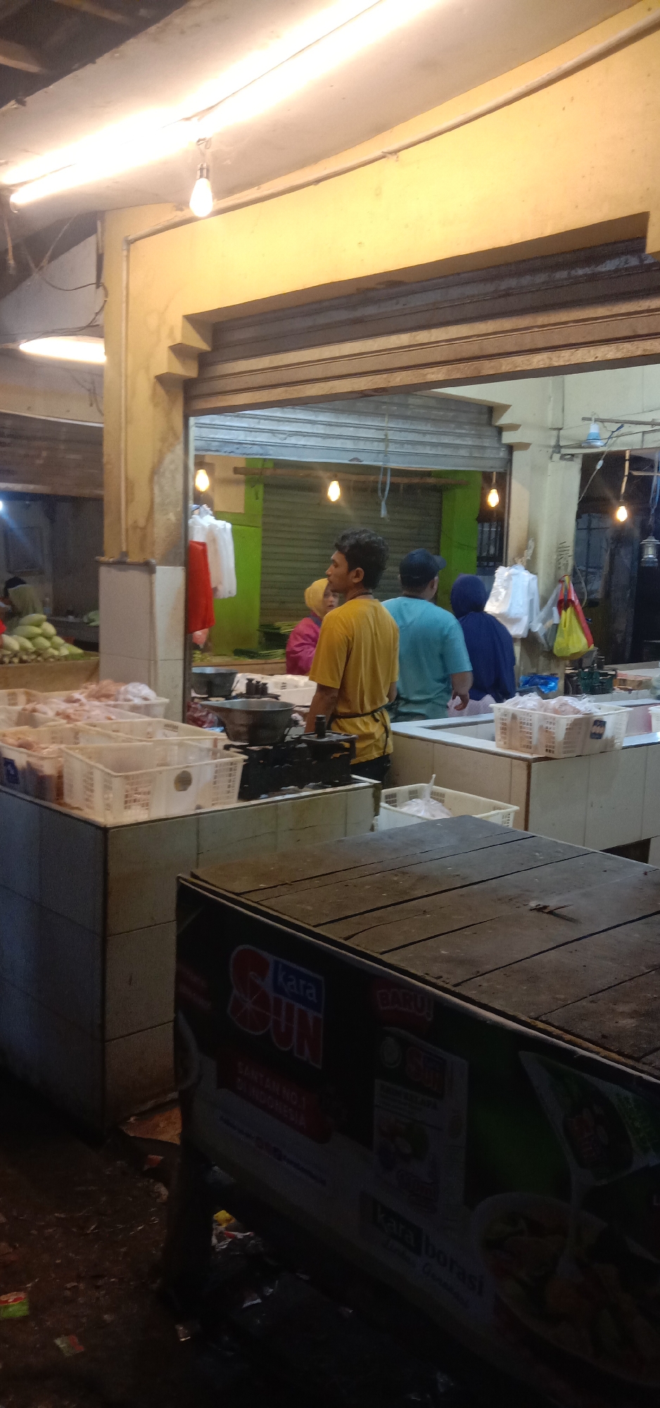 A man in a yellow shirt with an apron stands in a market in Subang, Indonesia. He is facing away from the camera, looking towards a counter with a black scale and several white plastic bins filled with raw chicken. Behind him, two other individuals are visible, one in a turquoise shirt and another in a blue headscarf. The market appears to be an open-air or covered stall with overhead lighting. In the foreground, a wooden table with a sign displaying "KARA SUN" is present. The environment is cluttered with some debris on the ground.