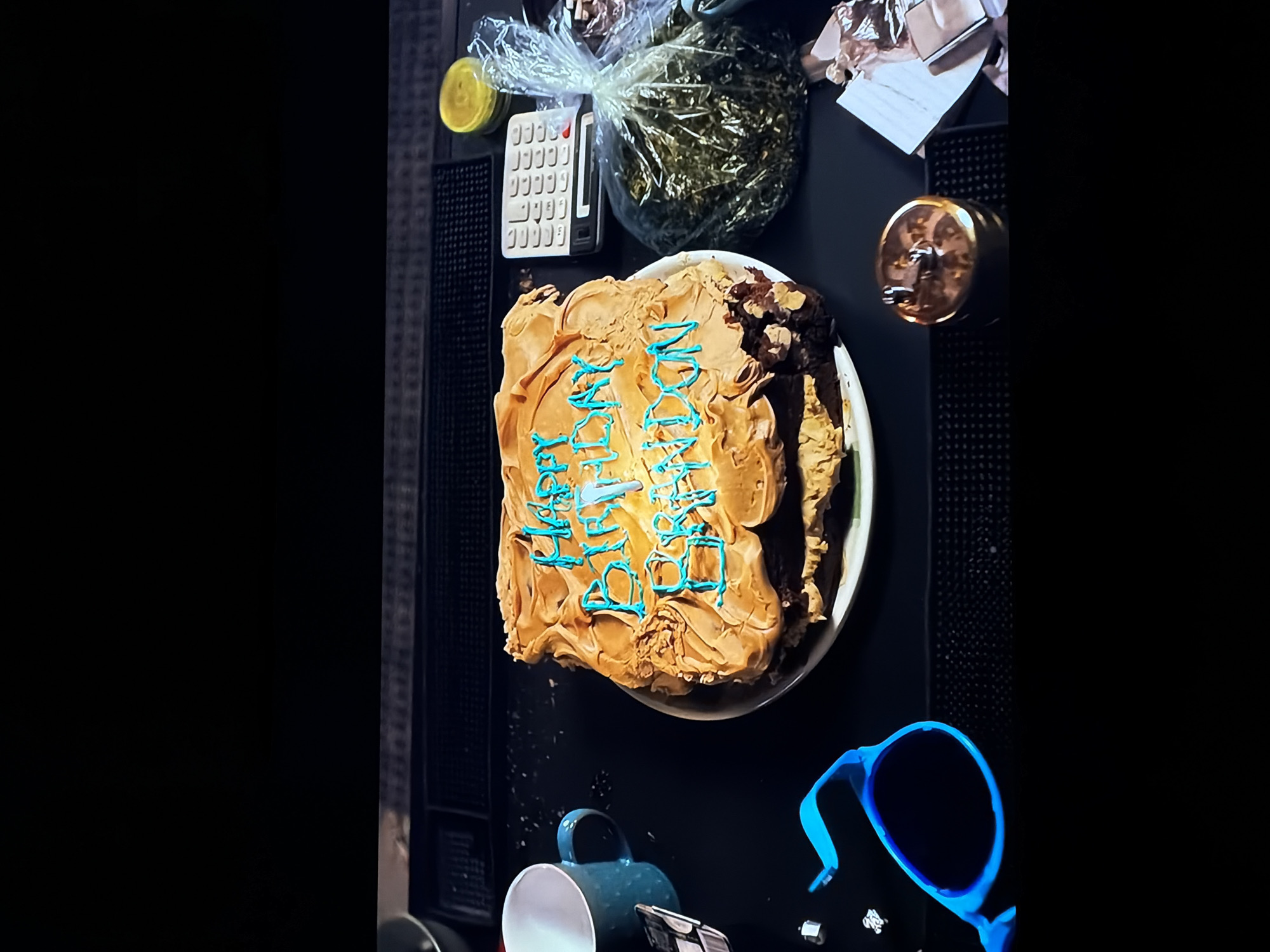 An overhead view captures a casual indoor setting, likely a table or countertop, in Sofiivska Borshchahivka, Ukraine. The central item is a square or rectangular cake, frosted with a light brown or tan icing. Written across the cake in bright blue icing are the words "HAPPY BIRTHDAY," indicating a celebration. A portion of the cake on the right side appears cut or partially consumed.
Clustered around the cake are several other objects. To the upper left of the cake, a white, vintage-style calculator sits next to a clear plastic bag that is tied at the top and contains a substantial amount of a dark green, leafy, or granular substance. A small yellow container or lid is also visible nearby. On the upper right, a metallic, cylindrical object with a golden sheen, possibly a grinder or a decorative container, is present. In the lower part of the frame, a teal-colored ceramic mug lies on its side, and a pair of vibrant blue sunglasses is placed to its right. Various other indistinct items, such as foil and paper, are scattered around the periphery. The dark surface of the table suggests an evening or indoor setting with artificial lighting. The overall scene suggests a low-key birthday celebration.