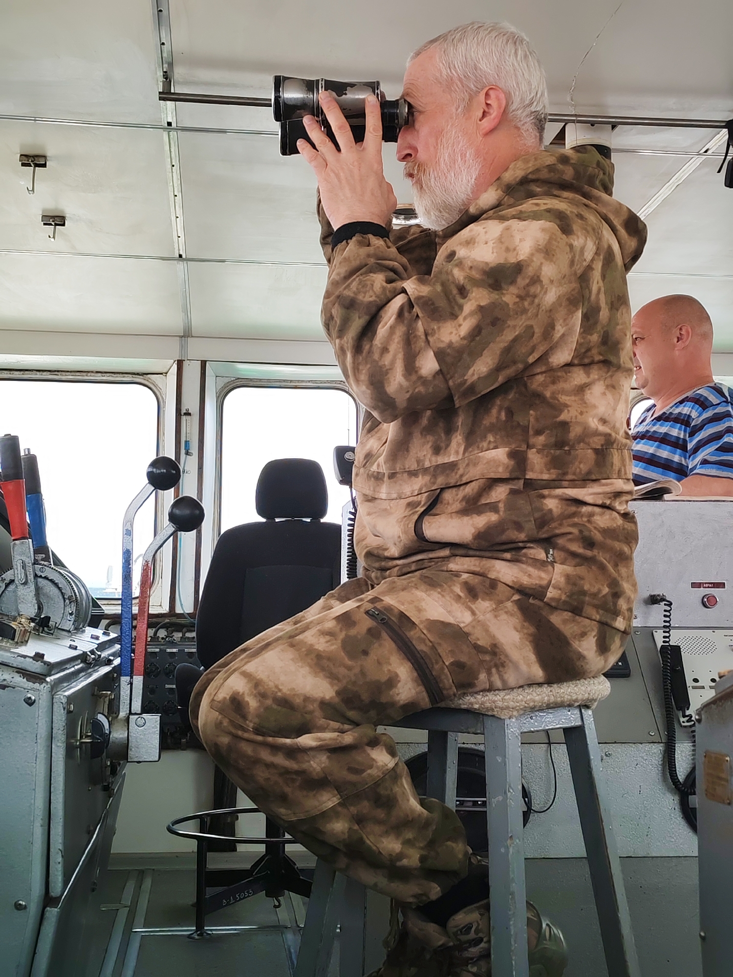 The image captures the interior of what appears to be a ship's bridge or control room, likely in Medvezhyegorsk, Russia. The main subject is an older man, seen in profile, with a white beard and grey hair, intently looking through a pair of binoculars. He is wearing a camouflage-patterned hooded jacket and matching trousers, seated on a high grey stool topped with a beige knitted cushion. His hands are up, holding the binoculars to his eyes.
In the background, to the right and partially obscured by the first man, another man is visible. He is bald with a light beard, wearing a blue and white striped t-shirt, and is looking towards the front of the vessel.
The control room is brightly lit by natural light streaming through large windows. To the left, there are several ship controls, including a complex panel with various levers featuring red, blue, and black handles. A black-seated chair is positioned behind these controls. The ceiling is white with metal structural elements and two small metal hooks.
On a console located to the right of the main subject, a coiled cable, a black microphone, and a red button are visible. Faint Cyrillic text is also present, possibly including "АВАРИЯ" (Emergency) near the red button. On the base of the stool, the alphanumeric code "B-L 5053" can be seen. The scene suggests an active watch or navigation during daylight hours.