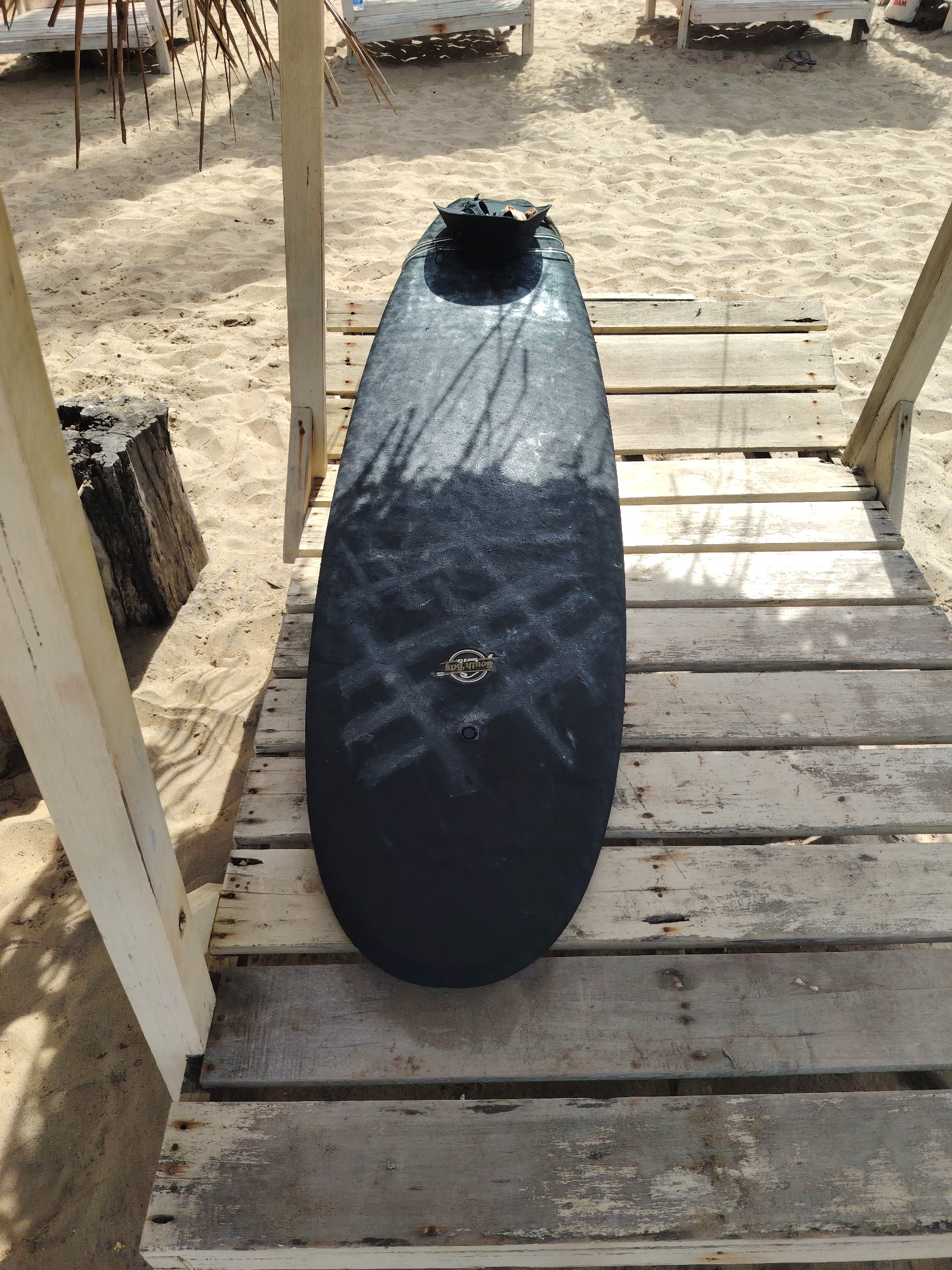 An overhead, medium shot captures a black surfboard with a textured surface placed on a weathered, light-colored wooden deck. The surfboard is positioned vertically in the frame, with its tail facing the top. A small, black bag is attached to the tail of the surfboard. The wooden deck is constructed from planks of varying widths and shows signs of age and wear, with some planks appearing cracked or discolored. The deck is situated on a sandy beach, with sand visible beneath and around it. In the background, white lounge chairs and palm fronds suggest a beach resort or recreational area. Sunlight casts dappled shadows across the surfboard and the sand, indicating it is a bright, sunny day.