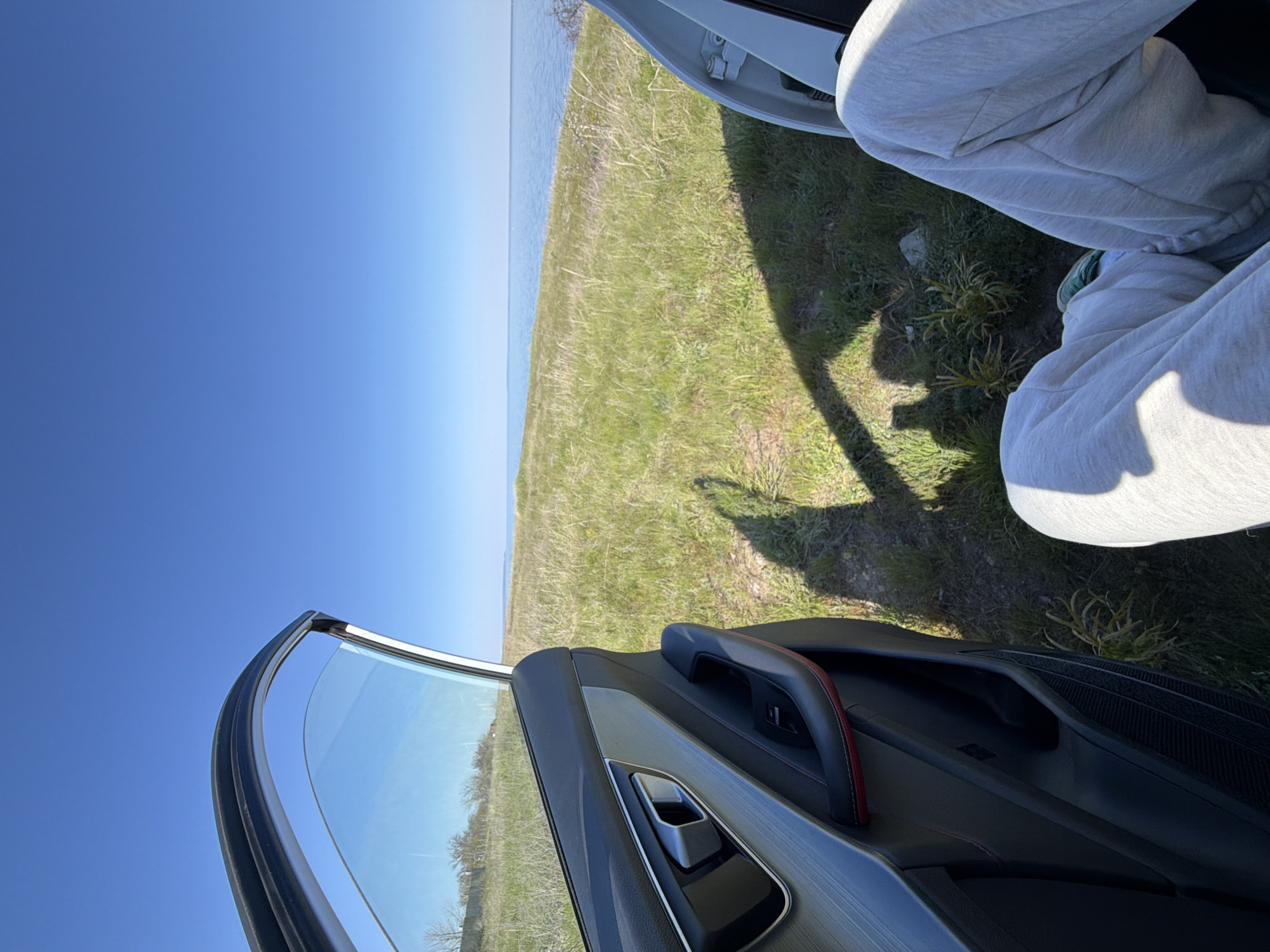 The image captures a scene on the outskirts of Kamyshin, Russia, near a body of water. The clear blue sky indicates a sunny day. An open car door frames the landscape, with a partial view of someone wearing light gray sweatpants sitting inside. The foreground shows the interior of the car door, featuring a black panel, a silver handle, and a red accent line. Outside the car, there is a grassy field leading to a water body, possibly the Volga River. The shadow cast by the car door and the person inside suggests the sun is positioned high in the sky.
