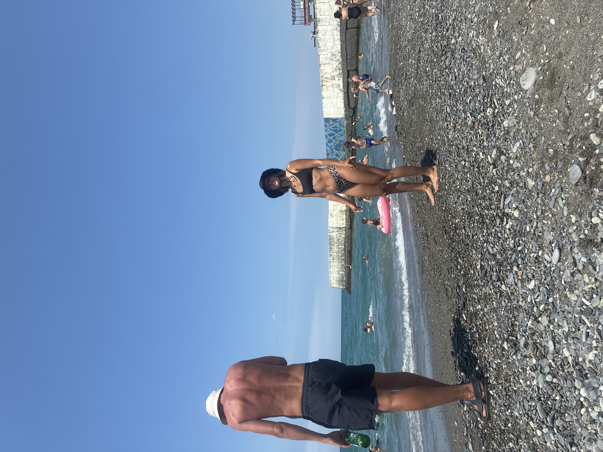 On a bright, sunny day at a pebble beach, likely in Sochi, Russia, two people are prominently featured. In the foreground, a man from behind wears a white bucket hat and black swim shorts. He holds a green bottle in his right hand. To his right, a woman stands facing forward, wearing a black bikini with a leopard print bottom and a black hat. Her body is tanned, and she wears a necklace. Behind them, the turquoise sea is filled with people swimming and playing. A pink inflatable ring floats in the water. In the background, a concrete structure, possibly a breakwater or pier, extends into the sea. The sky is clear blue.