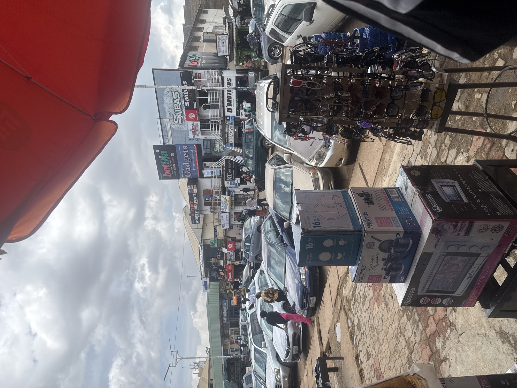 The image depicts a bustling street scene in Jos, Nigeria, likely a market or commercial area. The perspective is from a low angle, possibly looking up from a seated position, with a large, bright orange umbrella or awning dominating the top left of the frame, suggesting it's daytime and potentially sunny, though the sky is partly cloudy.
The scene is filled with various objects and people. In the foreground, a rack of sunglasses is prominently displayed for sale, with rows of different styles and colors. Behind this, a variety of cars are parked along the street, suggesting a busy thoroughfare. Some cars are white, others are darker colors like gray and blue.
The background features several buildings with shopfronts, indicating a commercial district. Visible signs offer clues to the businesses present. We can see names like "SAIBAS CLOTH," "bet9ja," "Gadgets," "LIT KIT SPORTS STORE & PRINTS," "O'BROTHERS," and "GADGETS & TRENDS." There are also signs for "Okey Dubai" and "VIN-THEO PLAZA." Boxes with images of kitchen appliances, specifically "MAXIMUS Cooking Appliances" and an "ELECTRIC TOASTER OVEN" from "MASTER CHEF," are stacked in the lower right, suggesting retail sales.
Several people are visible, some interacting with the stalls or vehicles. One person is seen wearing a yellow and black patterned shirt, and another is in a pink outfit. The general atmosphere appears to be one of active commerce and daily life. The paving stones of the street are visible, adding to the sense of a pedestrian-friendly or mixed-use area. The overall impression is a lively and vibrant street market.