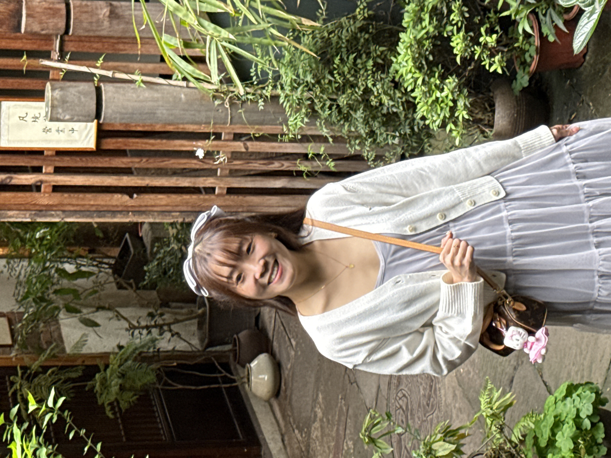 A female subject is depicted standing outdoors, smiling directly at the viewer. She has brown hair with bangs and wears a white and black striped headband. Her attire consists of a light grey tiered dress layered with a white knitted cardigan. A delicate gold-colored necklace with a small pendant is visible. She carries a small, brown patterned shoulder bag, from which a white and pink Hello Kitty keychain hangs. The ground beneath her is paved with irregular stone slabs, some displaying faint decorative patterns. The background features a blend of traditional architecture and lush greenery. To the left, potted plants and a dark entryway are visible. To the right, a wooden slatted fence or screen is prominent, adorned with a framed vertical sign displaying Chinese characters, which translate to "Fán jìng yíng chá zhōng" (Mortal Realm Tea House operating). Numerous potted plants, including bamboo-like stalks, are arranged around the wooden structure and a cylindrical wooden post. The setting is consistent with a traditional courtyard or pathway, likely within a cultural or commercial area of Yuzhong District, China, specifically a tea house environment.