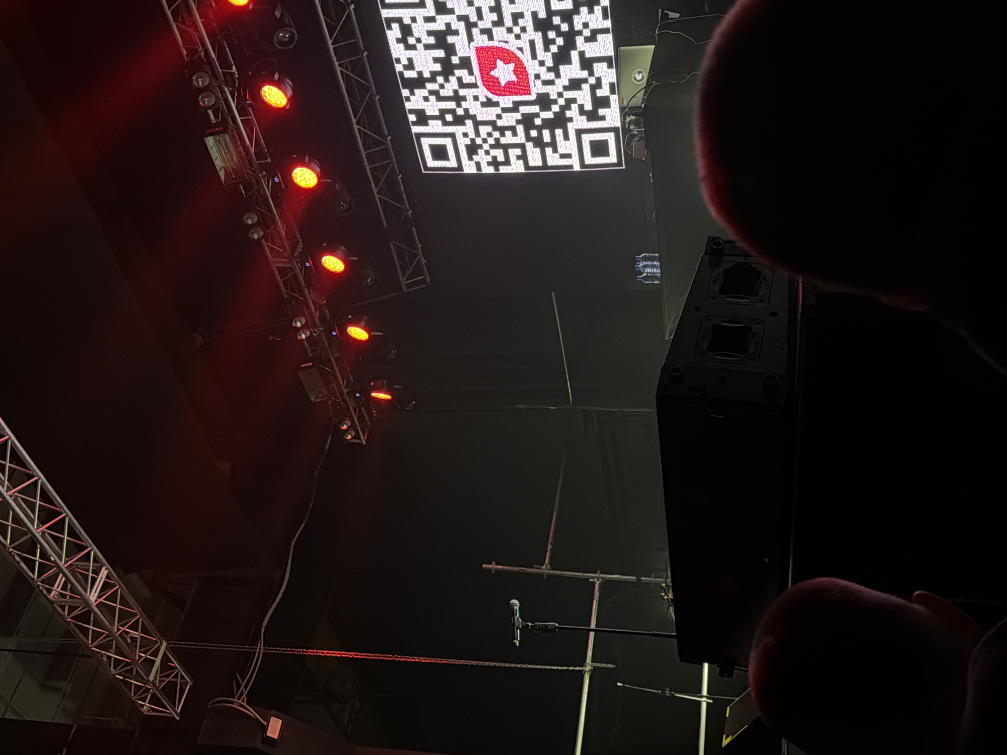This low-light photograph captures an upward perspective within an indoor event venue or concert hall in Perm, Russia, viewed from a close, possibly audience or backstage, vantage point.
The scene is dominated by stage equipment and lighting against a dark background. On the left, a metal truss structure supports a series of bright red and orange stage lights, casting a warm illumination downwards. These lights are a prominent feature, suggesting an active event or performance.
In the upper central portion of the image, a large display screen prominently features a bright white QR code. Embedded within the center of this QR code is a distinct red logo, which appears to be a stylized star or a multi-petal design.
To the right, a large, dark speaker system or sound equipment stack is visible. The immediate foreground on the right is obscured by an out-of-focus, dark hand or fingers, contributing to a first-person perspective.
Further within the frame, a microphone on a stand can be seen, positioned as if ready for a speaker or performer. Additional metal support structures and a horizontal red chain or cable traverse the dark space. The overall atmosphere is dark and dramatic, typical of a live event setting.