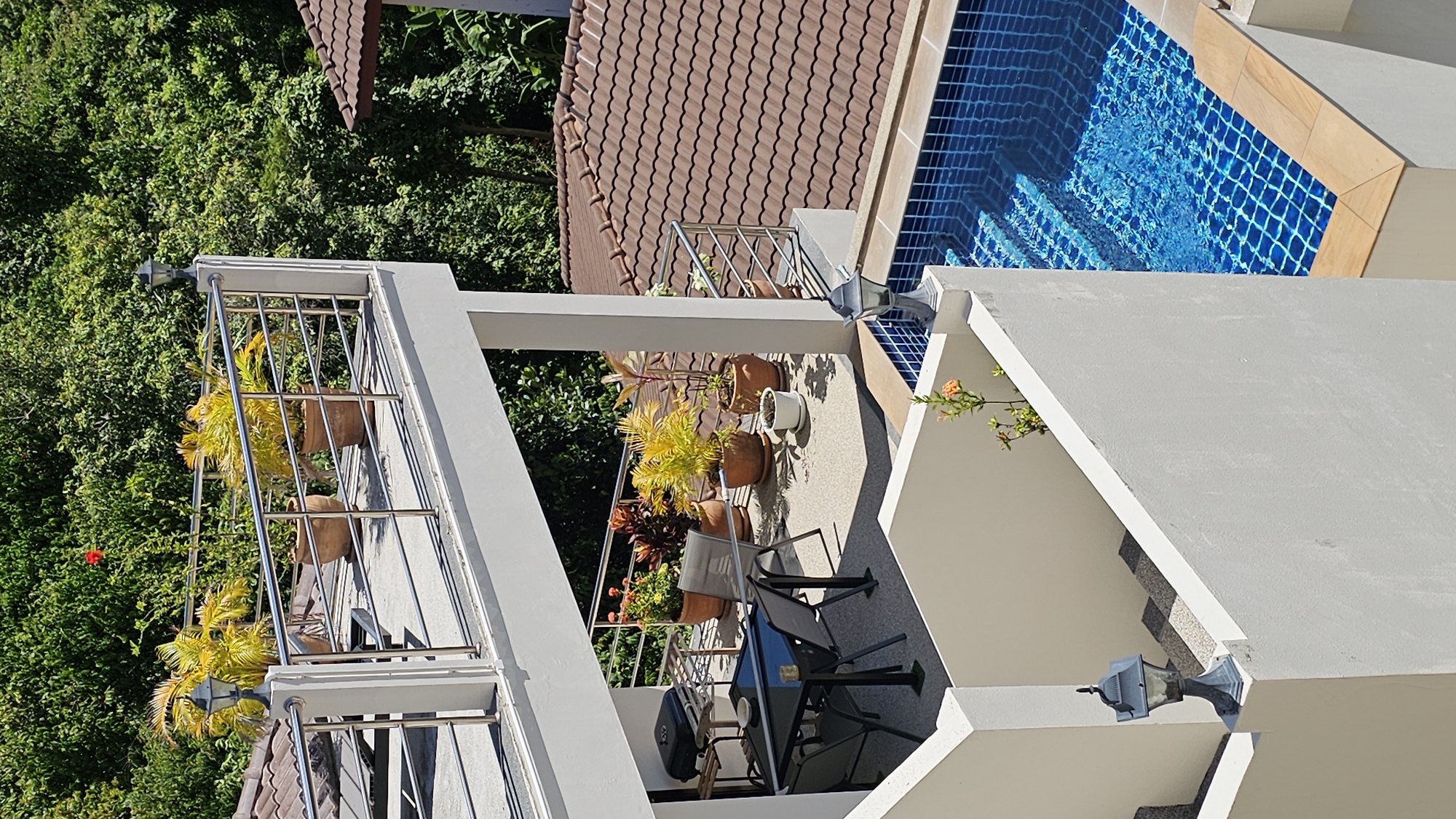 This is a high-angle, medium shot of an outdoor entertainment area with a swimming pool at a property in Baan Chaweng, Thailand. A covered patio with a dining set and a barbecue grill is visible in the mid-ground. Potted plants are arranged on the patio and on an upper-level balcony. The swimming pool, tiled in blue, is in the foreground and right of the frame. The scene is surrounded by lush green foliage.