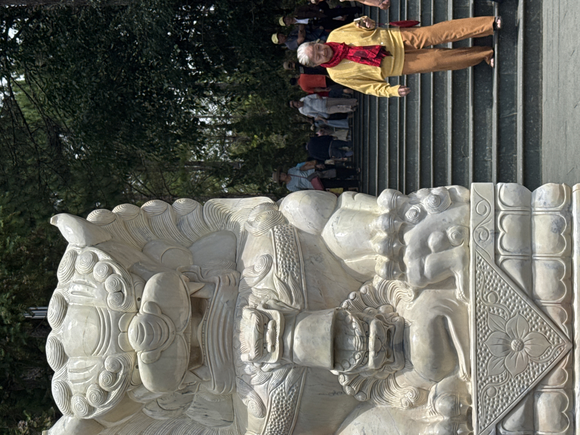 The image shows a large, ornate marble statue of a lion in the foreground, with a group of people walking up a wide stone staircase behind it. The statue is intricately carved with swirling patterns and sharp teeth, suggesting an Eastern architectural style. The people on the stairs are dressed in casual clothing, and some are looking towards the statue. The background consists of lush green trees, indicating an outdoor setting. This statue is located at the entrance to the Linh Phuoc Pagoda in Da Lat, Vietnam, a prominent Buddhist temple known for its elaborate mosaics and architecture.