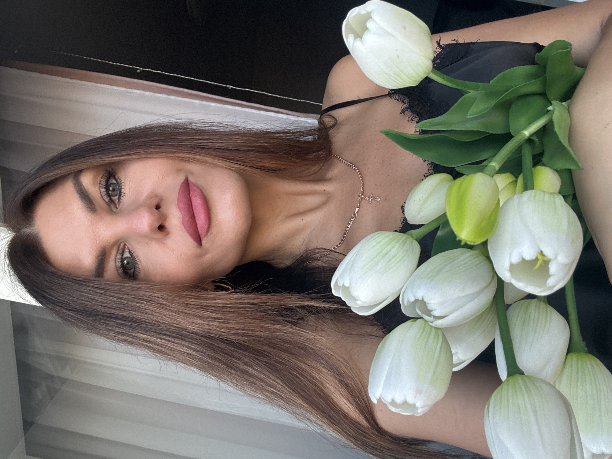 A woman is holding a bouquet of white tulips. She is wearing a black top with lace trim and a gold necklace with a cross pendant. She has brown hair and is wearing pink lipstick. She appears to be indoors, near a window. I am unable to determine the exact location, or if the photograph was taken in Chaikovsky, Russia.