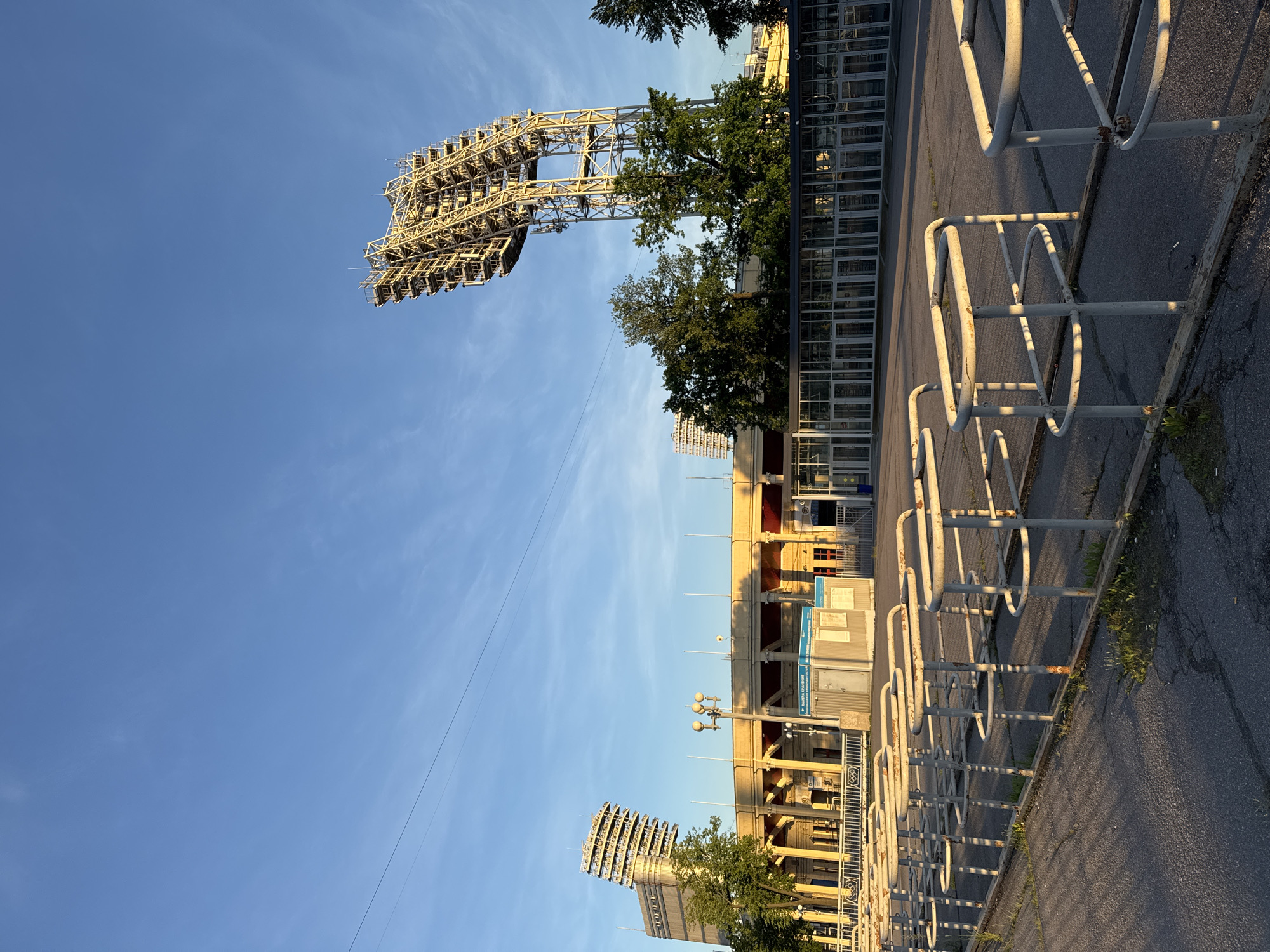 This is a photograph of a stadium in Saint Petersburg, Russia. The image captures an outdoor view of the stadium's exterior on a clear, sunny day. In the foreground, there are rows of metal turnstiles, suggesting an entrance area for spectators. The turnstiles are white and appear somewhat weathered. Beyond the turnstiles is a paved area, and further back is the stadium building itself. The stadium has a distinctive architectural style with a series of columns supporting an upper structure. Two large, metal floodlight towers are prominent features, one on the left side and a larger one on the right side of the image. These towers are tall and skeletal in construction. The sky is a bright blue with a few wispy clouds. Trees with lush green foliage are visible behind the stadium building and around the floodlight towers. The sunlight casts long shadows across the scene, indicating it might be early morning or late afternoon. There are no people visible in the image. The overall impression is one of a large sports or event venue in a city setting.
