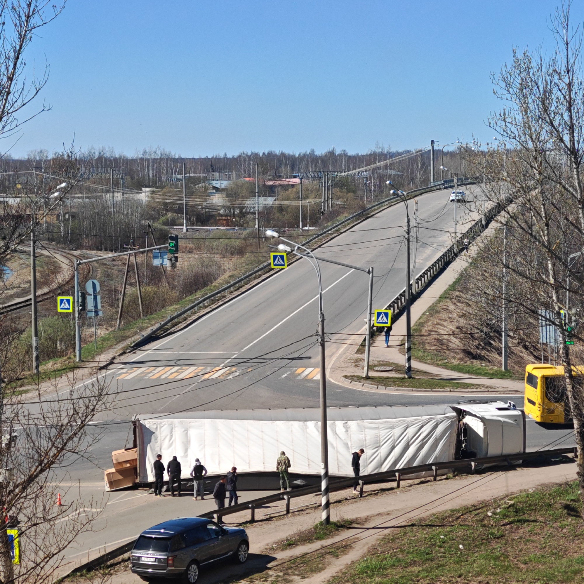 The image shows a road in Rostov, Russia, where a large white semi-truck has overturned, blocking part of the intersection. Several people are standing near the overturned truck, some appearing to assess the situation. To the left of the truck, there is a dark-colored SUV. A yellow bus is stopped on the road behind the truck. The road leads up an overpass with pedestrian crossing signs. The background features bare trees and a clear blue sky. Railway tracks and power lines are also visible in the background.