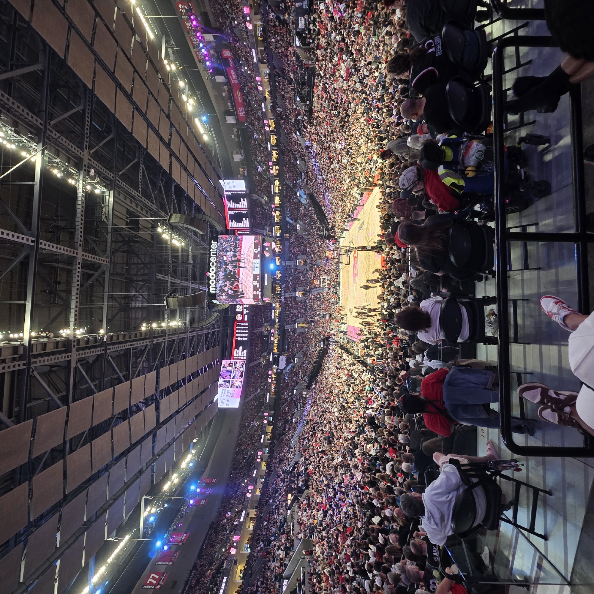 An aerial, wide-angle, full-shot captures a basketball game in progress at the Moda Center in Portland, Oregon, USA. The arena is filled with a large crowd of spectators, many of whom are wearing team colors of red and black. The basketball court is visible in the center of the image, with players on the court. The arena's ceiling features a complex structure of beams and lighting fixtures, with acoustical panels suspended from the ceiling. A large video scoreboard hangs from the center of the arena, displaying game information and video replays. The overall atmosphere appears to be energetic and festive, with the crowd actively engaged in the game.