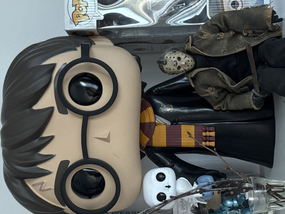 This image depicts an indoor display of various figures on a white surface, presumed to be a shelf or table, located within a structure in Moscow, Russia. The central and most prominent subject is a large, stylized vinyl figure, identifiable as Harry Potter due to its black hair, round black glasses, a distinct lightning bolt scar on the forehead, and a red and gold striped Gryffindor scarf worn over a black robe.
To the left of the large Harry Potter figure, a smaller, white stylized owl figure, resembling Hedwig, is positioned. In front of and slightly to the left of the owl, a small clear glass vessel holds multiple bare tree branches. To the right of the Harry Potter figure, a smaller, articulated action figure, identifiable as Jason Voorhees, is seated. This figure is characterized by a white hockey mask with red circular markings, a distressed brown jacket over a black shirt, dark pants, and a dark machete-like weapon held in its right hand. In the background, to the far right, a partially visible clear-boxed Funko Pop! figure, displaying the "Pop!" logo, indicates a collector's item within the display.