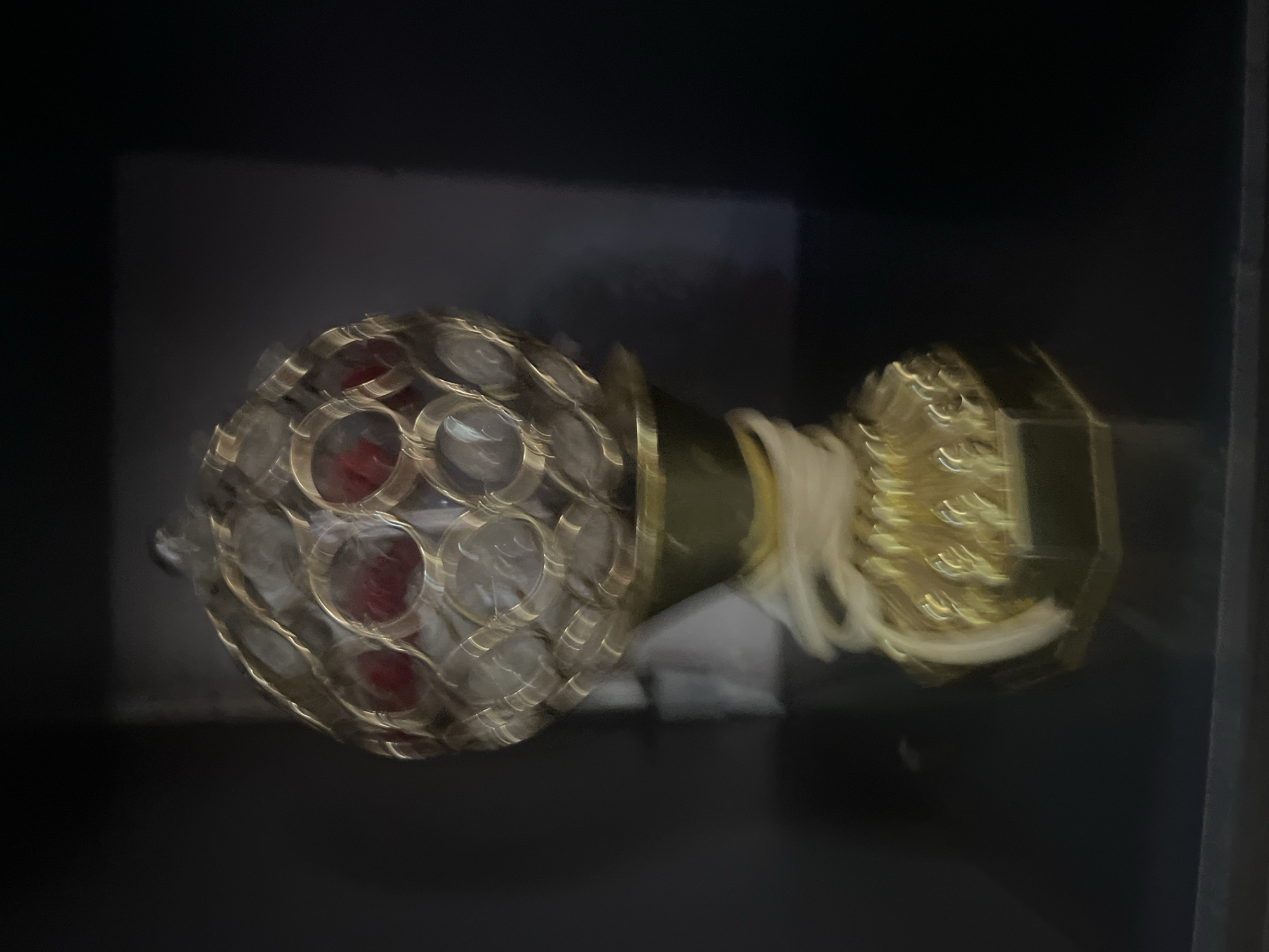 A decorative object, possibly a trophy or ornamental lamp, is displayed against a dark background, likely a shelf or cabinet. The object has a hexagonal gold-colored base with an intricate embossed pattern. Rising from the base is a golden stem wrapped with a light-colored ribbon-like material. At the top, a spherical cage-like structure is made of gold-toned metal, encasing what appear to be clear or translucent decorative elements and some circular red inserts. The overall image is slightly blurred, with the focus primarily on the object. There is no indication of the city of Jos, Nigeria, or any other specific location in the provided image.