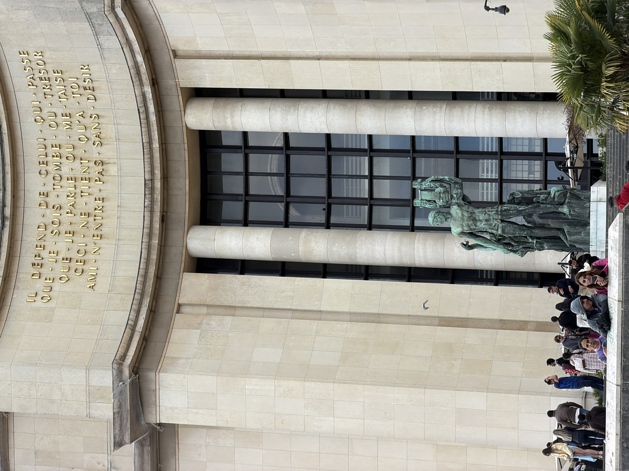 The image shows a view of the Palais de Chaillot in Paris, France. A crowd of people is gathered along the edge, likely overlooking the Jardins du Trocadéro and the Eiffel Tower. A bronze sculpture stands in front of the building's façade, which features large windows and stone columns. Above the columns, an inscription in gold lettering is visible on the building. A palm tree is on the right. A security camera is visible near the top.