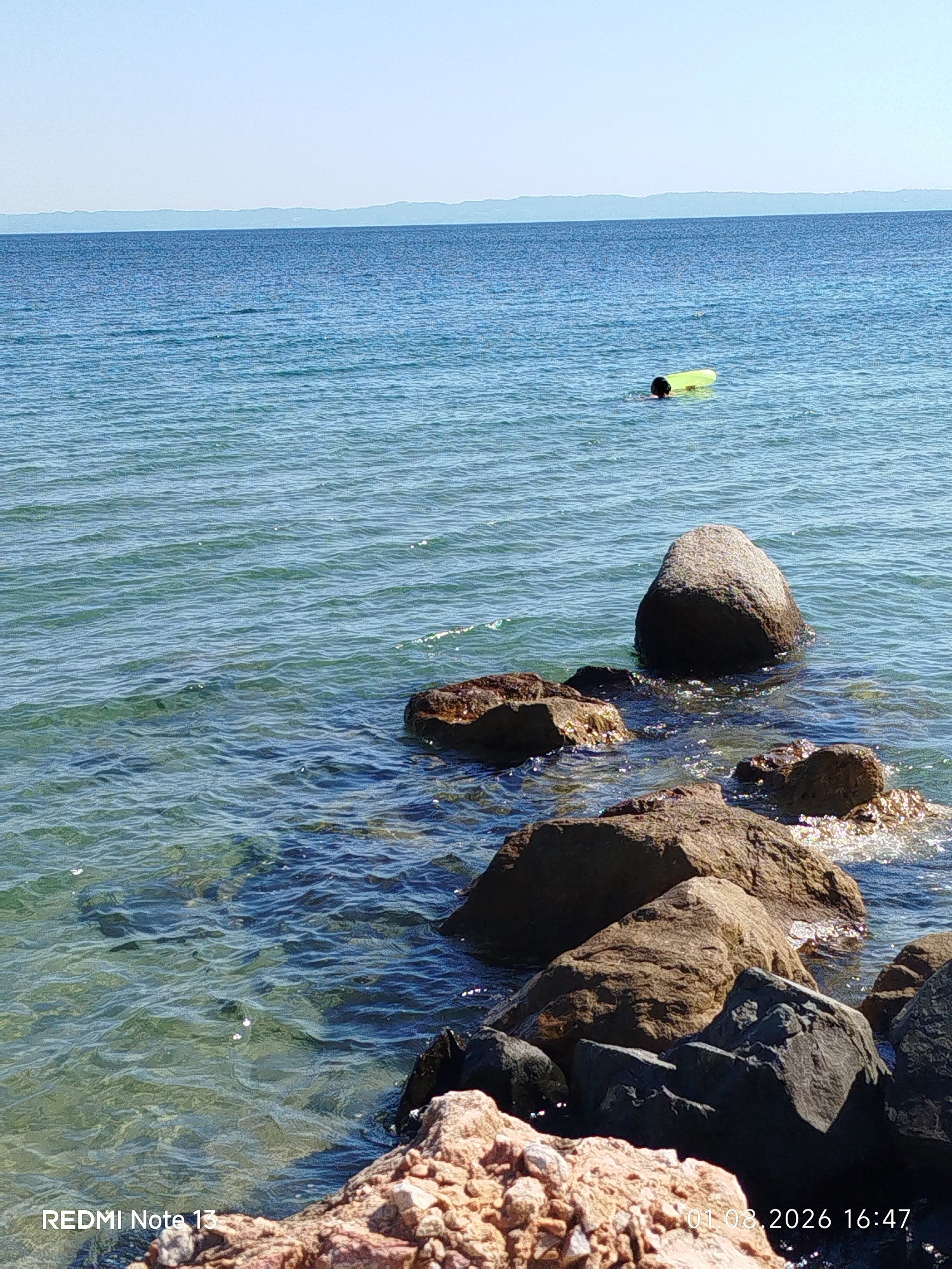 A clear, sunny day is depicted with a view of the Aegean Sea in Sithonia, Greece. The water is a vibrant turquoise and appears calm with gentle ripples. Several rocks, some partially submerged, are scattered along the shoreline in the foreground, with one larger, smooth, grey rock standing prominently in the mid-ground. In the distance, a yellow inflatable raft is visible floating on the water, with a person swimming near it. The horizon is marked by a hazy, distant coastline under a bright blue sky. The bottom left corner of the image shows the text "REDMI Note 13" and the bottom right displays the date and time "01.08.2026 16:47".