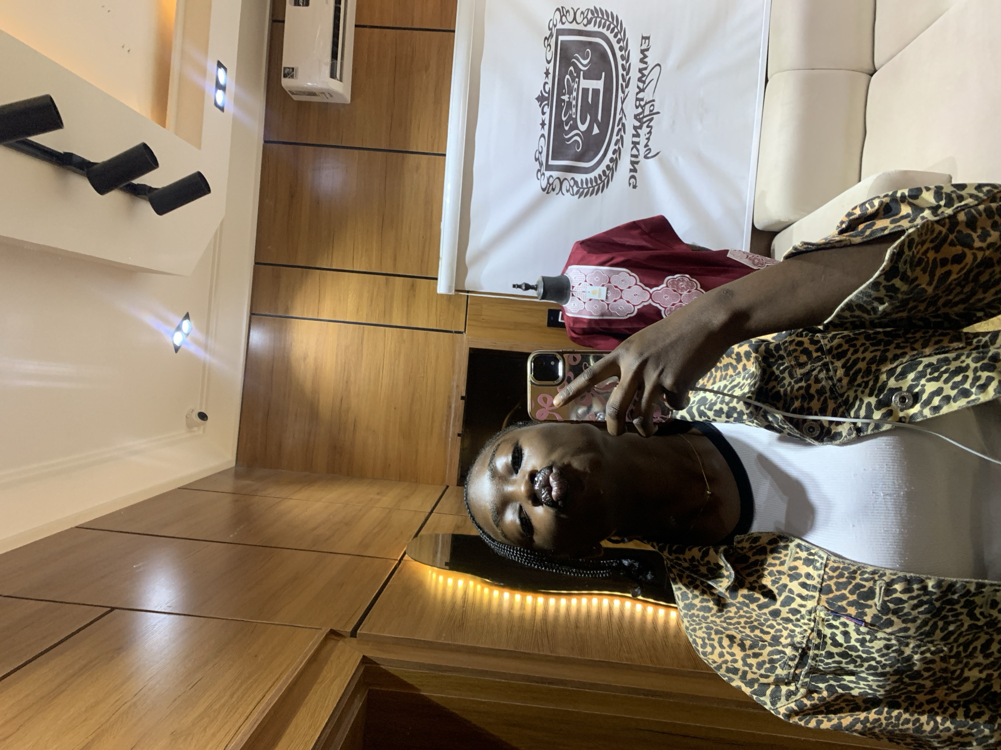 In Gura Top, Nigeria, a young Black woman with braided hair takes a selfie in a room with wooden paneling and modern lighting. She is wearing a leopard print jacket over a white top and has her lips pursed in a kissing gesture. In the background, a white banner displays a logo with the text "EMMYBANKING" and a crown emblem. A red garment is displayed on a mannequin beside the banner. A cream-colored sofa is visible to the right of the frame. The environment suggests a retail or display space, possibly a boutique or a showroom.