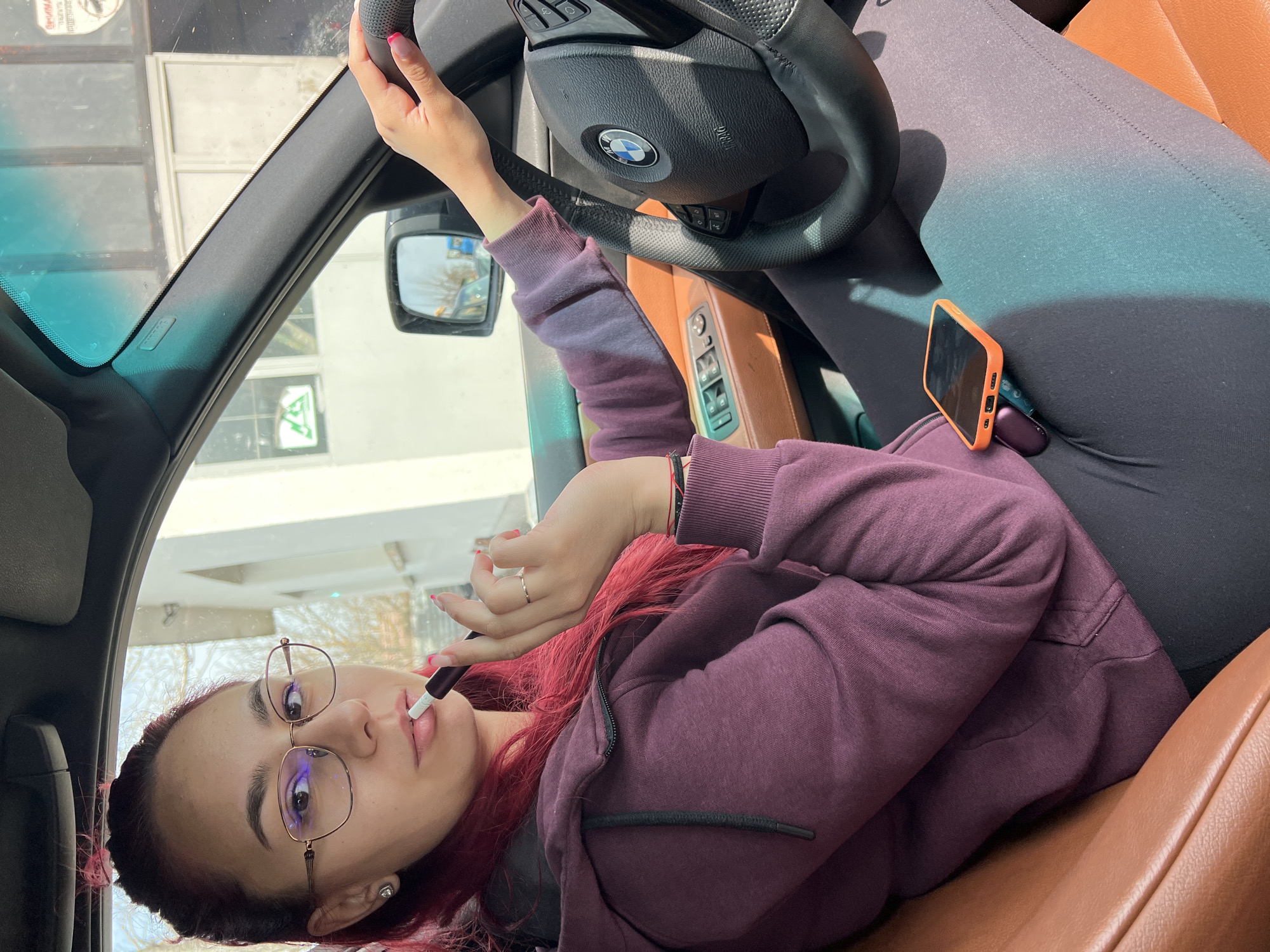 A young woman with pink-red hair and glasses is lying on the passenger seat of a BMW car. She is wearing a purple hoodie and grey leggings. She is holding an e-cigarette to her lips. An orange-cased phone is lying on her lap. The interior of the car has brown leather seats. The scene appears to be daytime based on the daylight visible through the car windows. There is no specific indication of Vabel, Bulgaria in the image.