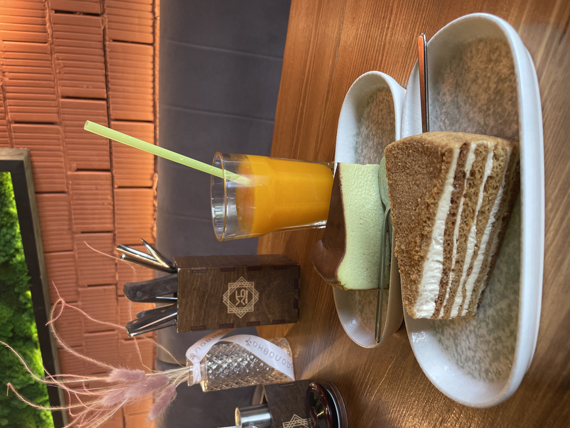 The image depicts a table setting at what appears to be a restaurant or cafe. A glass of orange juice with a yellow straw is placed next to a rectangular wooden holder containing cutlery. Near the cutlery holder, there is a small, ornate glass vase with decorative grass stems tied with a ribbon that has text in Russian written on it.
Two pieces of cake, each with a distinct color, are served on a narrow, curved white dish. A slice of light green and dark brown cake sits next to a piece of layered caramel cake with white icing. A metal spoon rests between the slices.
The table is wooden, and the background features a brick wall and a gray wall. A patch of green foliage is visible in the lower left corner. The scene gives the impression of a relaxed meal or snack setting. The Russian text on the ribbon suggests that the location is in Kazan, Russia.